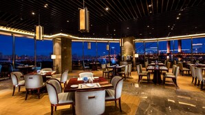 A spacious dining area with multiple tables and chairs, large windows offering a view of the cityscape at night, and a modern ceiling design. - Hilton Wuhan Riverside (Wuhan)