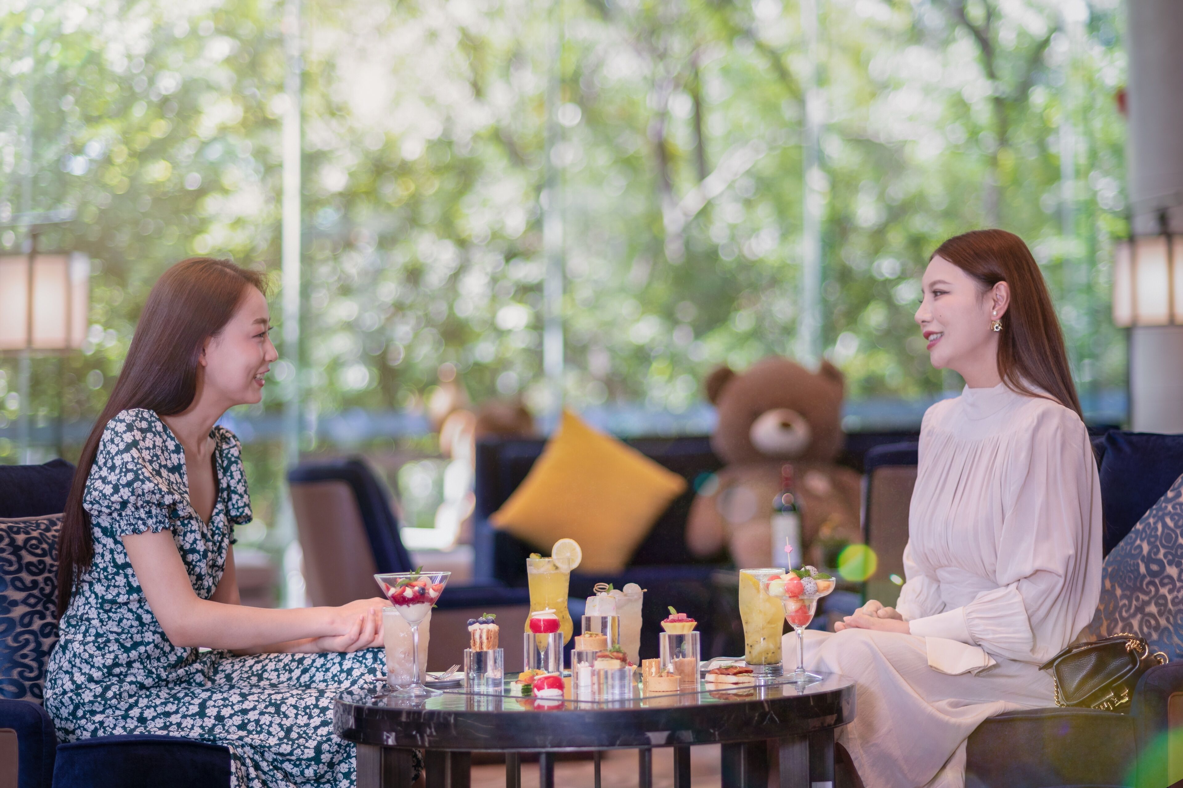 Two people seated at a round table with various desserts and drinks. The setting appears to be an outdoor lounge area with a view of trees and a teddy bear in the background.