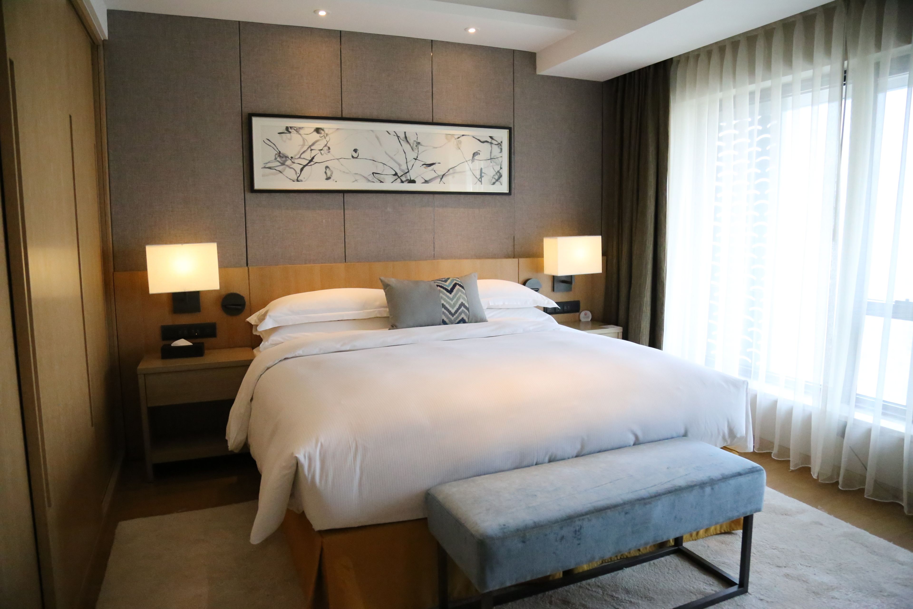Una habitación de hotel moderna con una cama grande, lámparas de noche, un banco, una obra de arte enmarcada en la pared y un ventanal con cortinas.