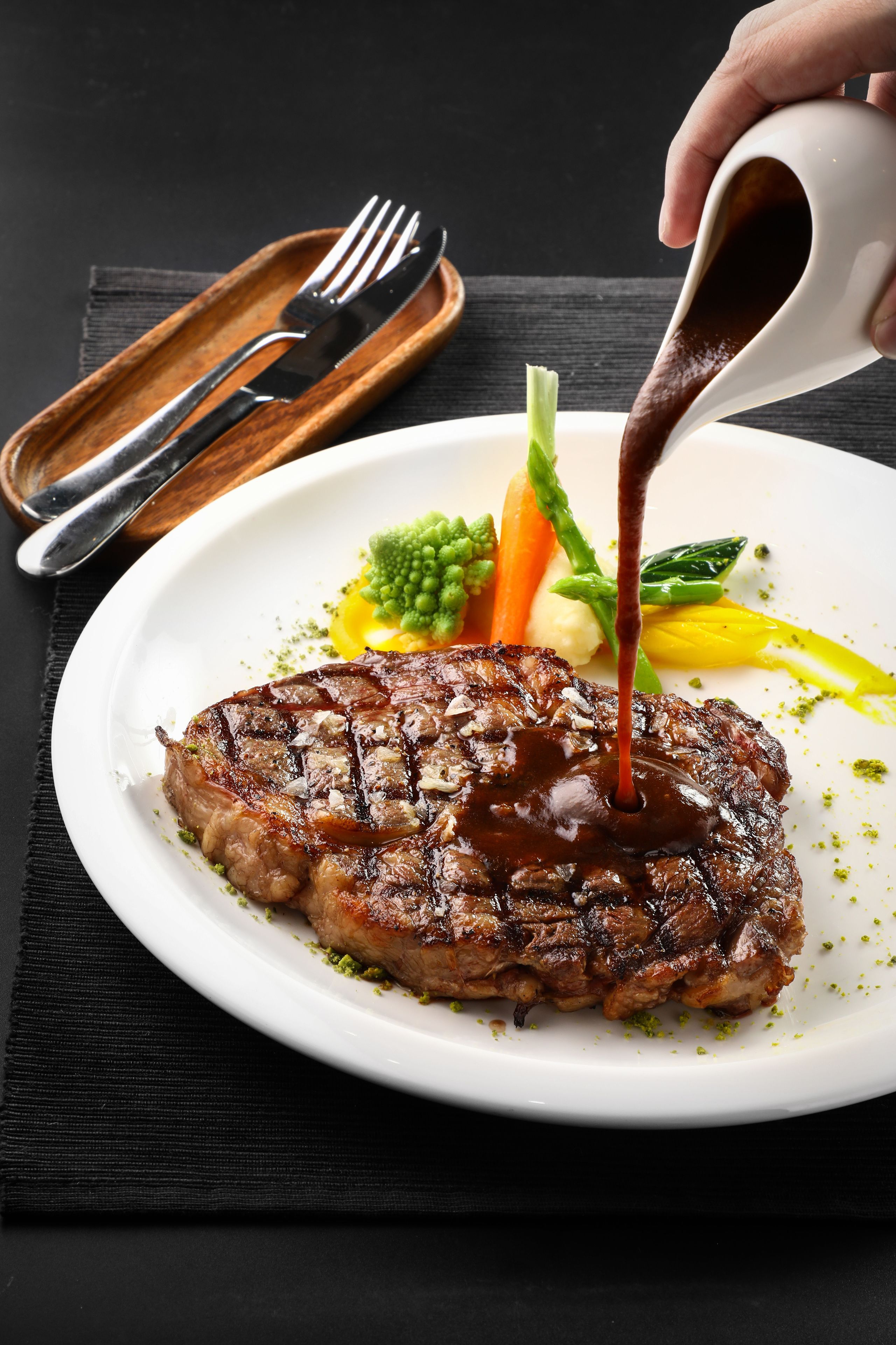 Ein Steak mit Soße auf einem Teller, dazu Gemüse und Besteck auf einem Tisch.