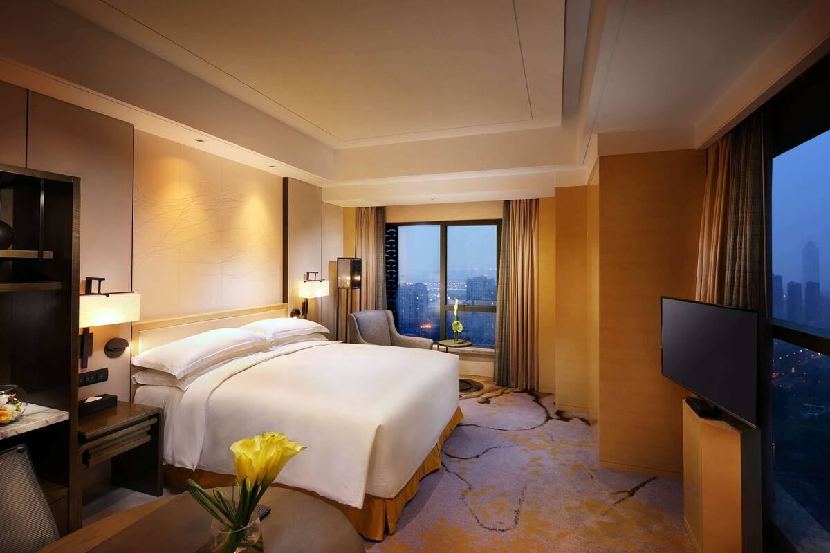 A hotel room with a large bed, a TV, a desk with a lamp, a chair, and a view of the city through a large window.
