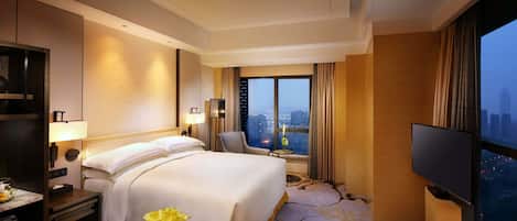 Habitación de hotel con una cama grande, televisión, escritorio con lámpara, silla y vistas a la ciudad a través de un ventanal amplio.