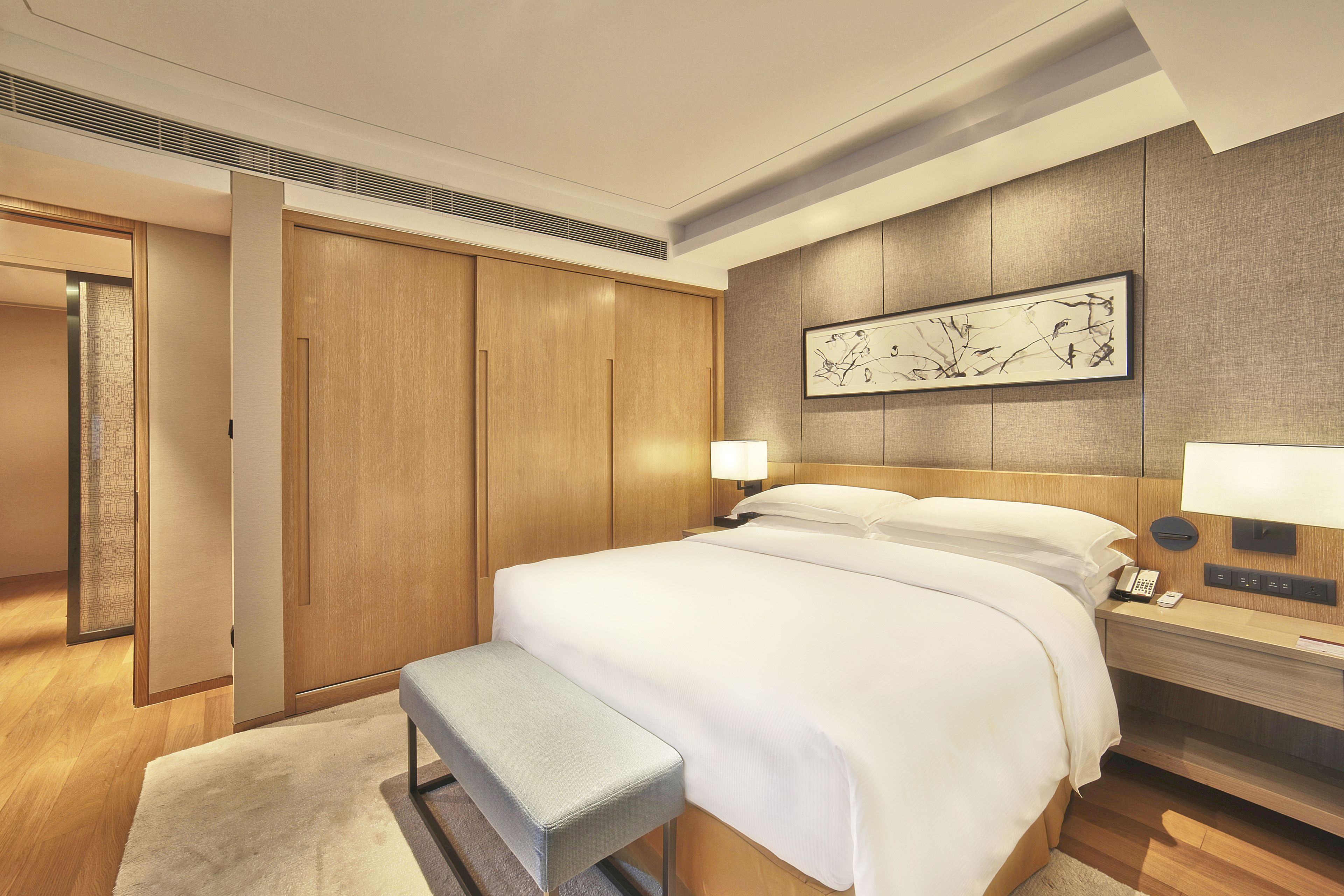 A modern hotel room with a large bed, wooden wardrobe, bedside table with lamp, and artwork on the wall.