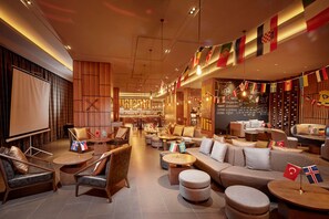 A spacious lounge area with multiple seating options, including sofas and armchairs, arranged around a central coffee table. Flags are displayed on the walls, and there is a chalkboard with writing on the back wall.