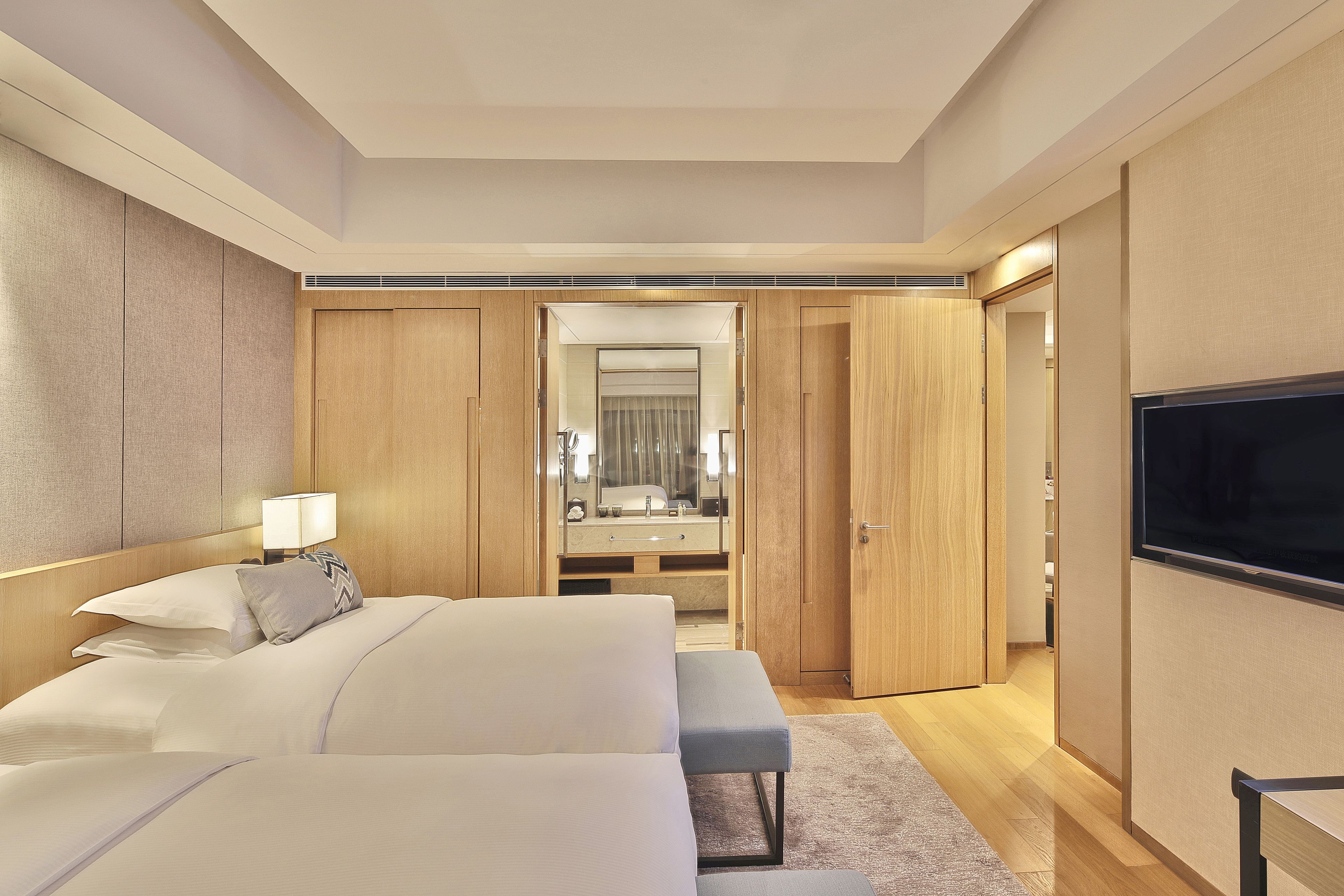 A modern hotel room with a large bed, a bench, a TV, a mirror, and a walk-in closet.