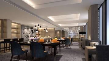 A modern hotel lobby with a long dining area, multiple tables, and chairs. There are pendant lights, a large abstract painting on the wall, and a patterned carpet.