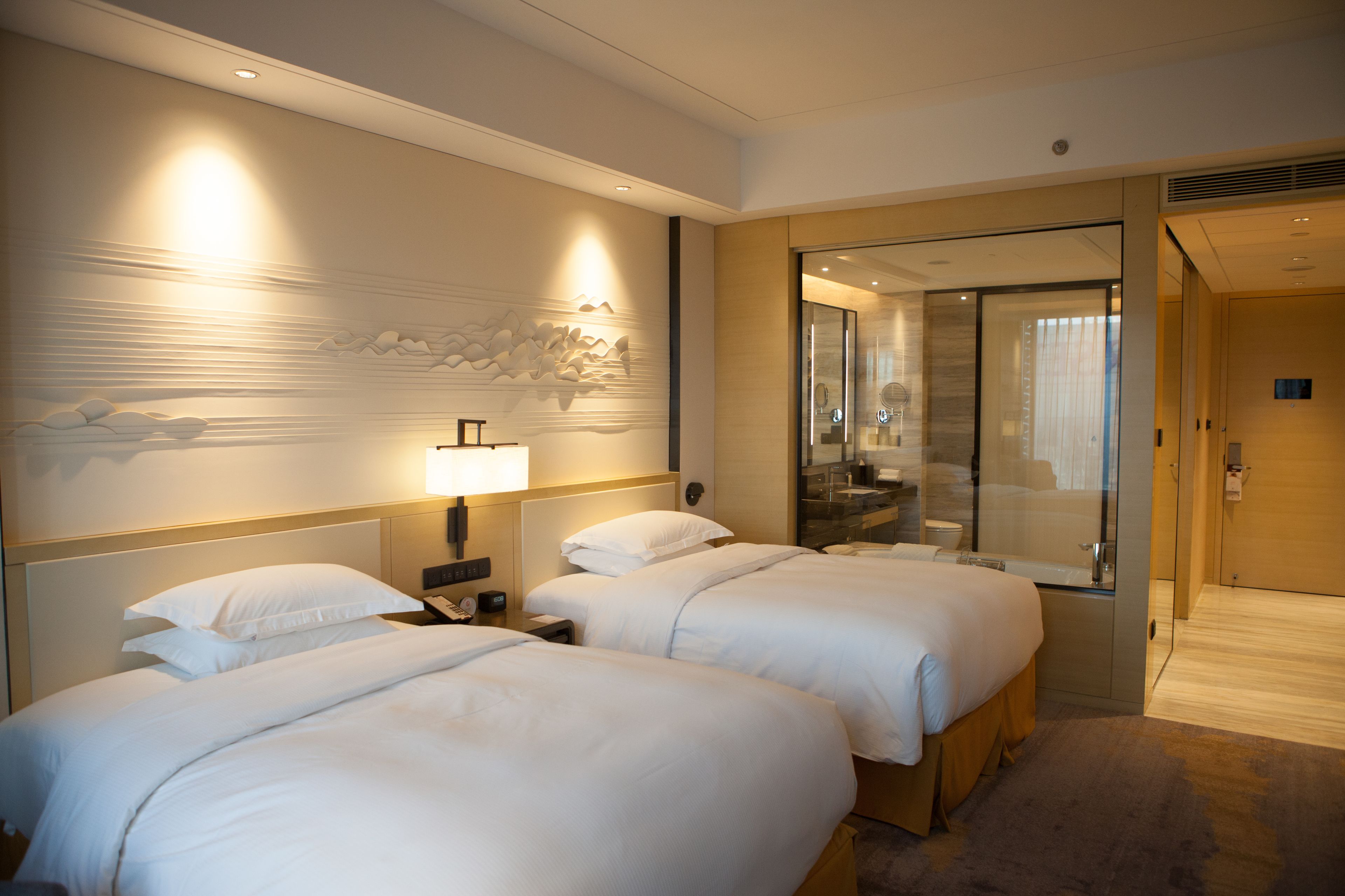 Ein Hotelzimmer mit zwei Betten, einem Nachttisch mit Lampe, einem Wandbild, einer Duschkabine aus Glas und einer Tür zu einem weiteren Zimmer.