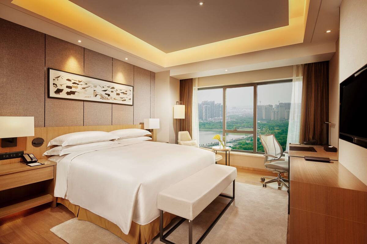 A modern hotel room with a large bed, a desk, a TV, and a view of the city.