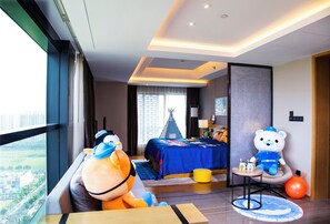 A hotel room with a large window, a bed with a blue bedspread, a teddy bear, a plush toy, and a small table with a lamp.