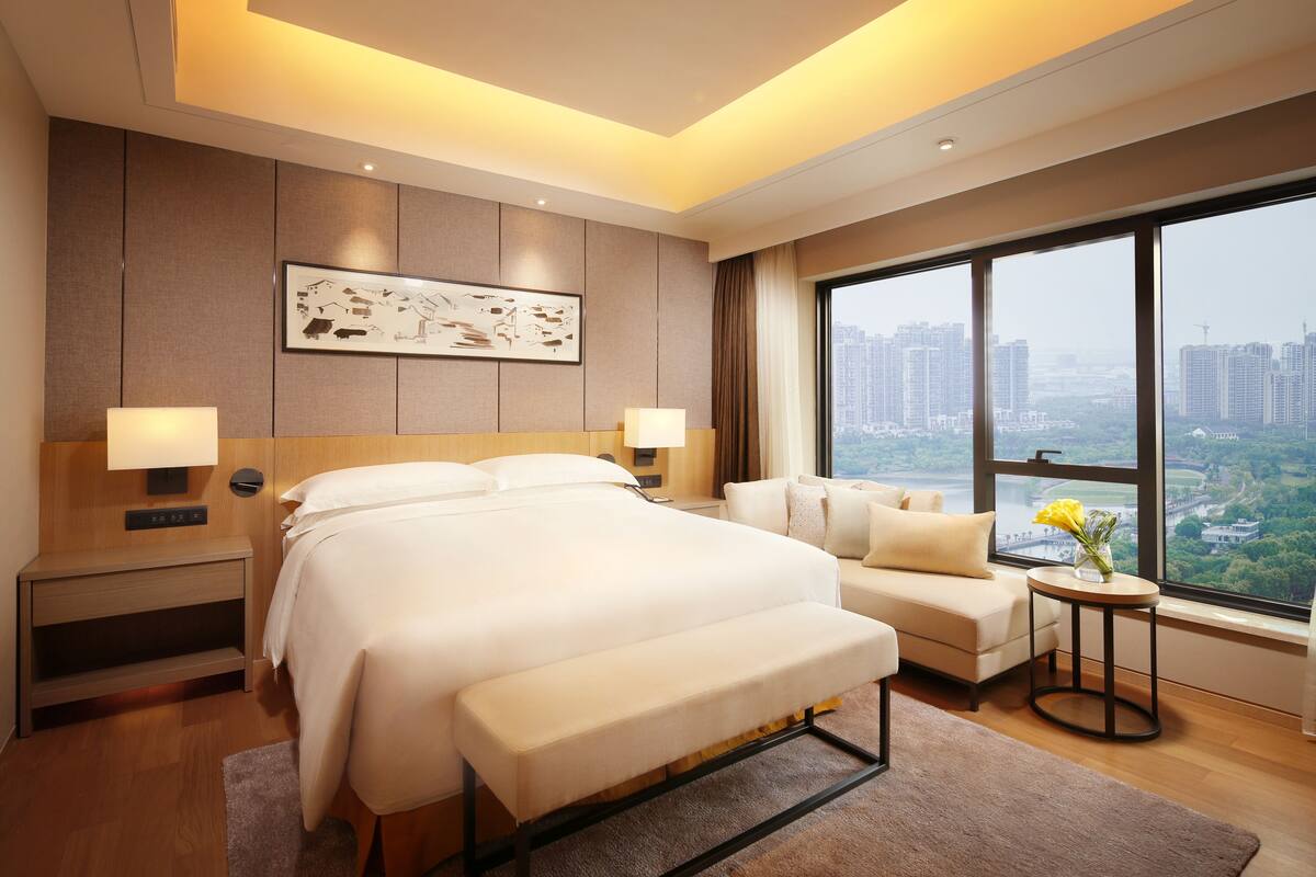 A hotel room with a large bed, a sofa, a small table with a vase, and a large window with a city view.