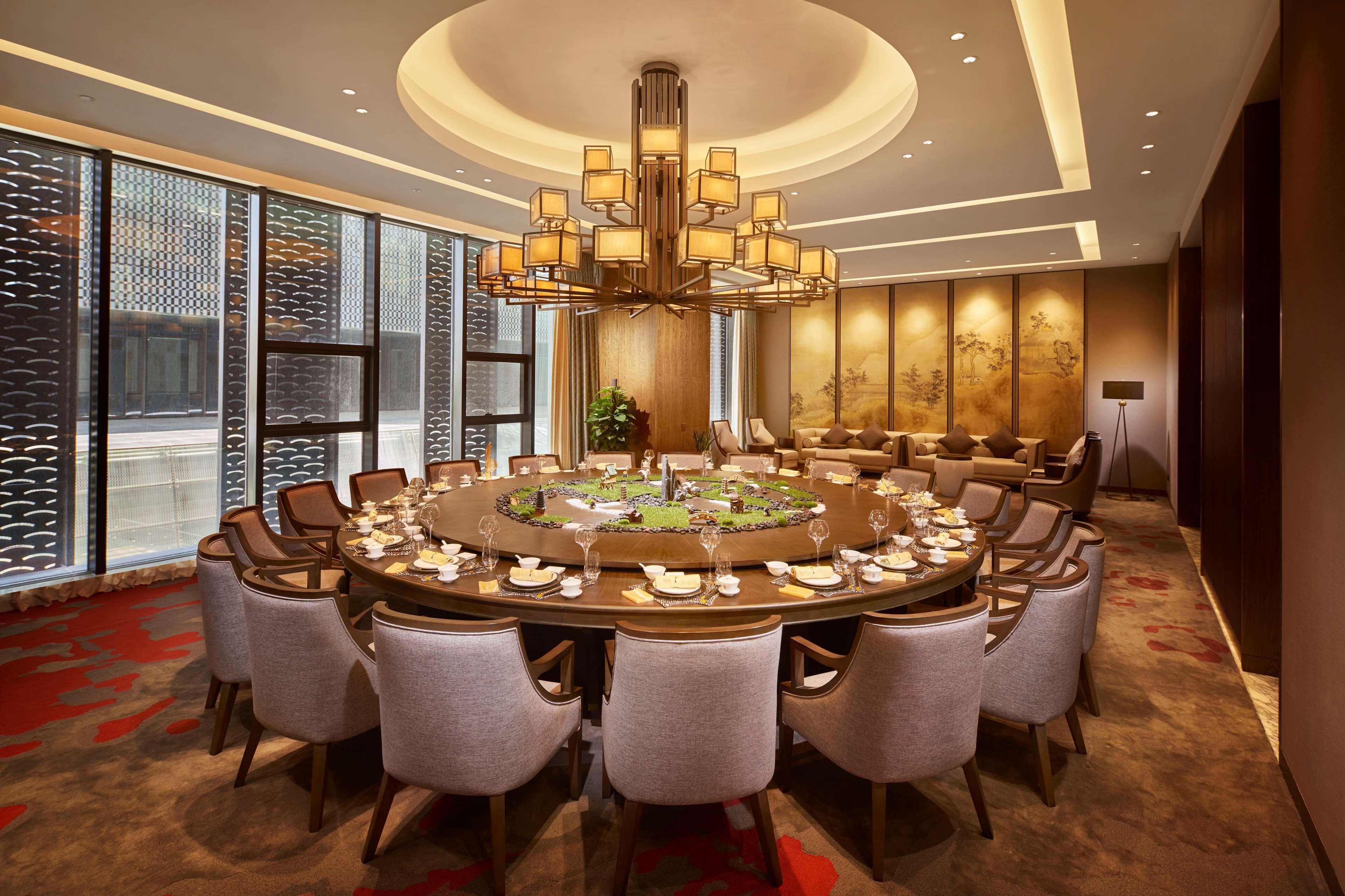 A round dining table set for a meal with multiple chairs, a chandelier, and a large window with patterned blinds.