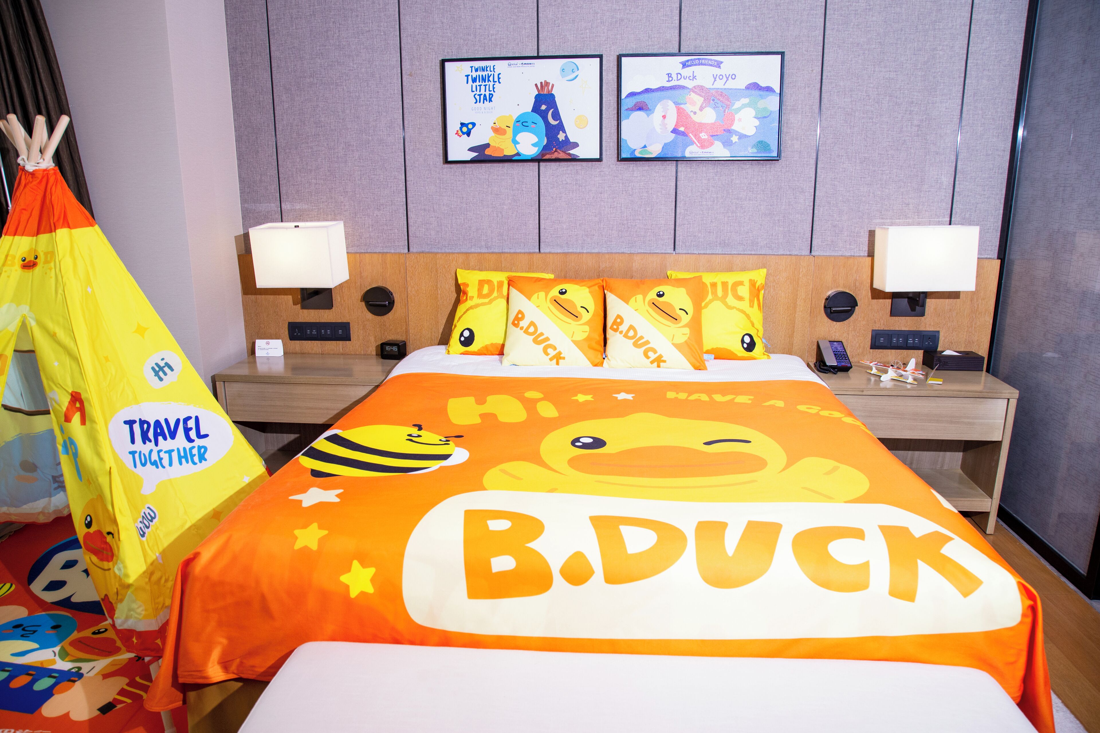 A hotel room with a bed featuring a B. Duck themed bedspread, a bedside table with a lamp, a wall-mounted clock, and a framed picture above the bed.