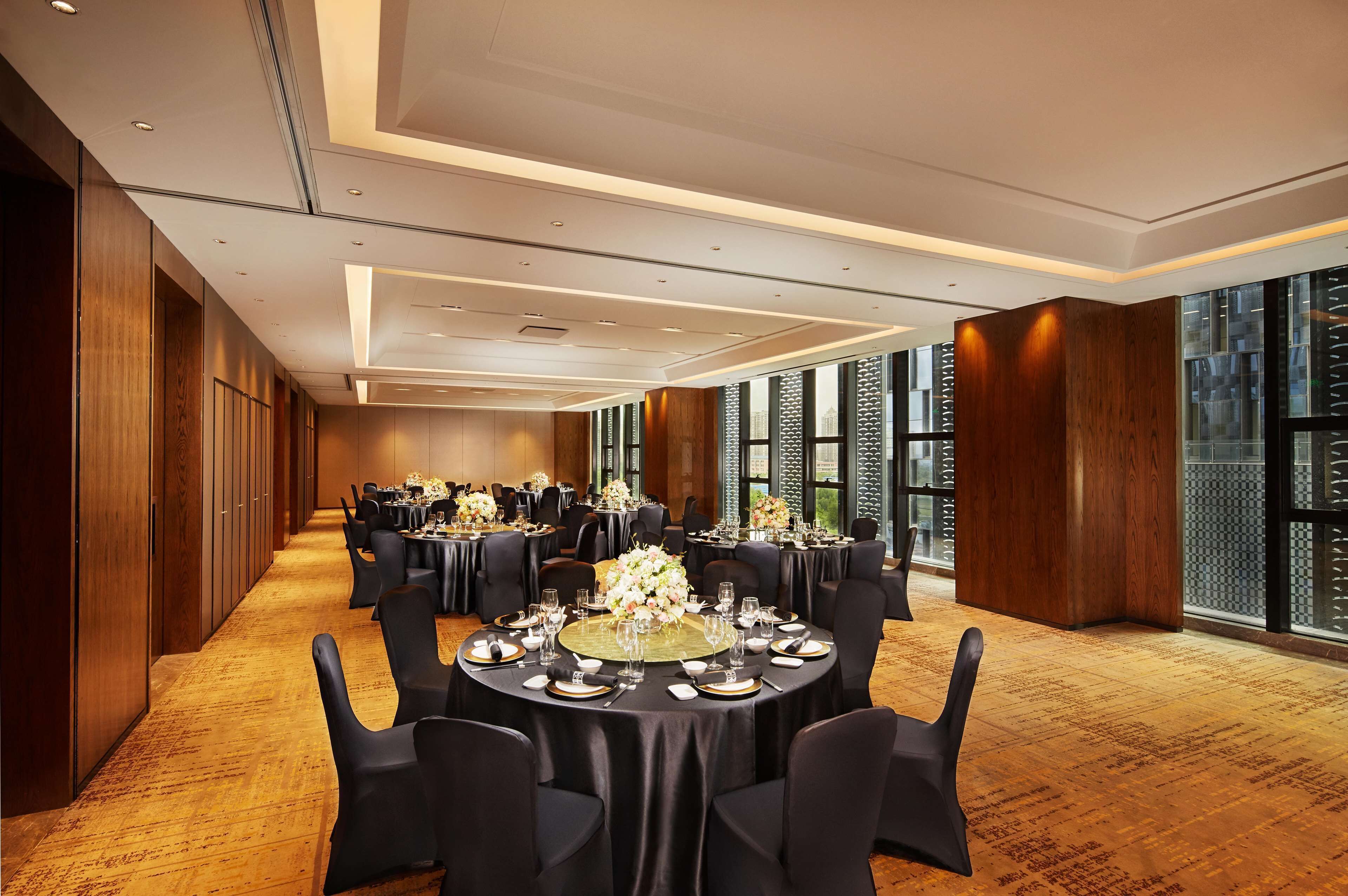 A spacious banquet hall with round tables set for dining, large windows offering views, and a wooden floor.