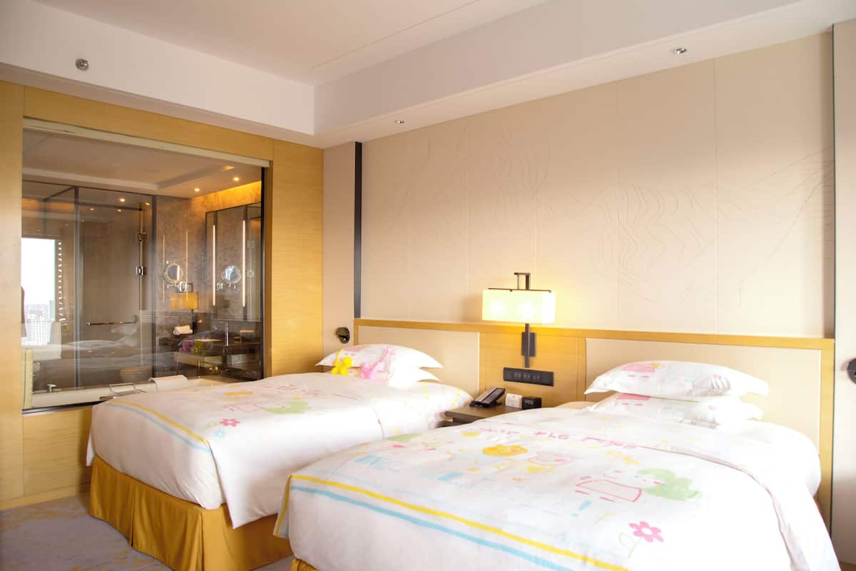 A hotel room with two beds, a large mirror, a bedside table with a lamp, and a view of the city through the window.
