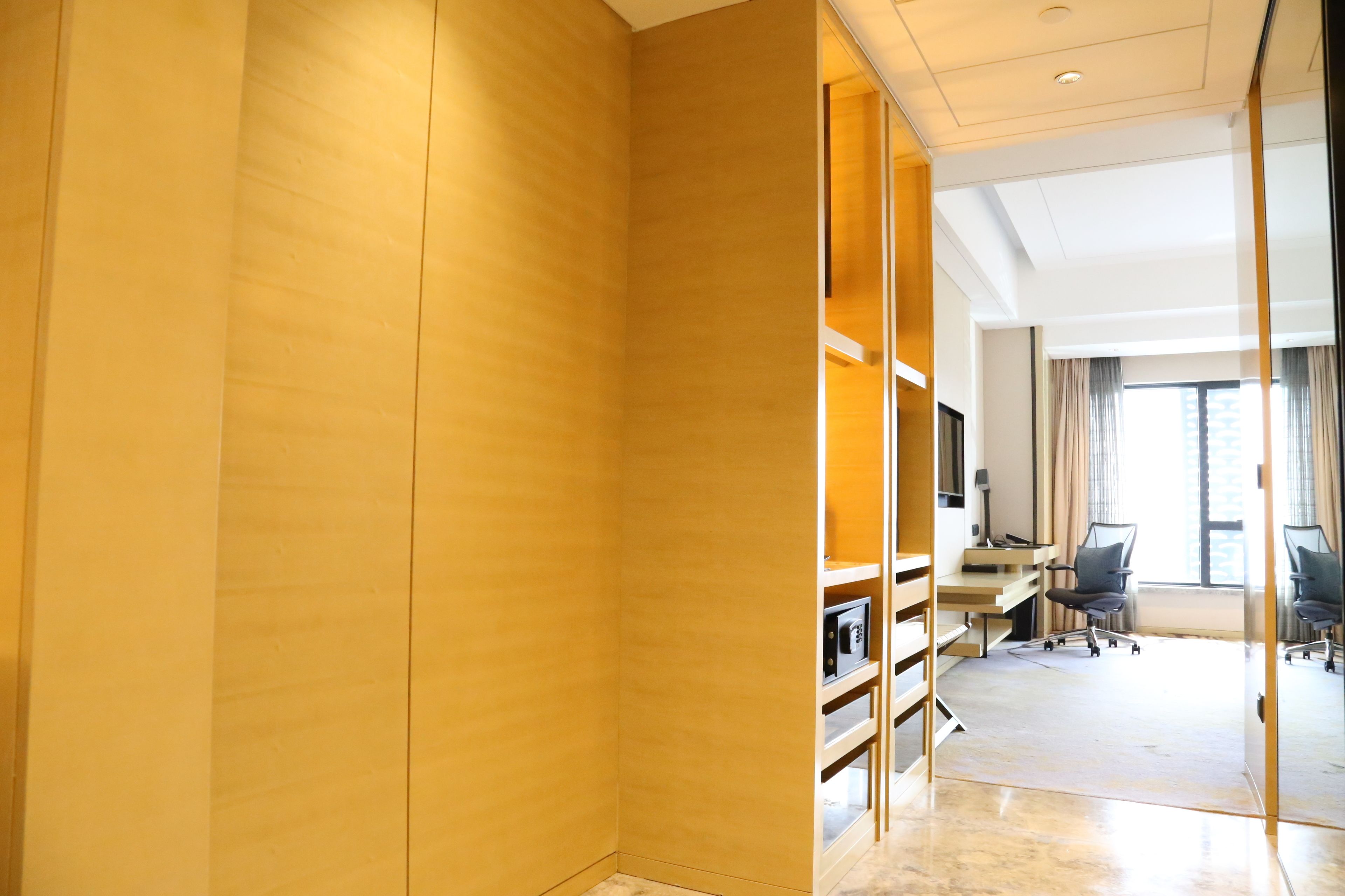 A modern hotel room with a wooden accent wall, a desk with a computer, a chair, and a large window with curtains.