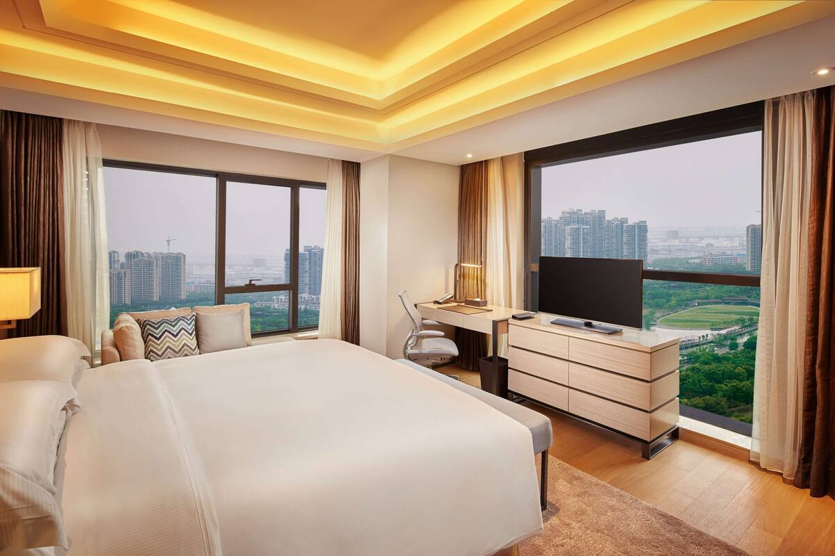 A hotel room with a large bed, a desk with a TV, and a view of the cityscape.