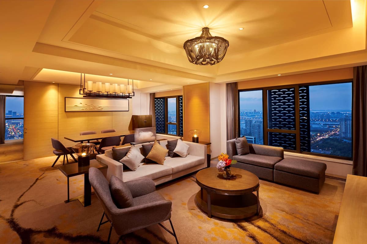 A modern hotel room with a large sofa, a dining table, and a view of the cityscape through large windows.