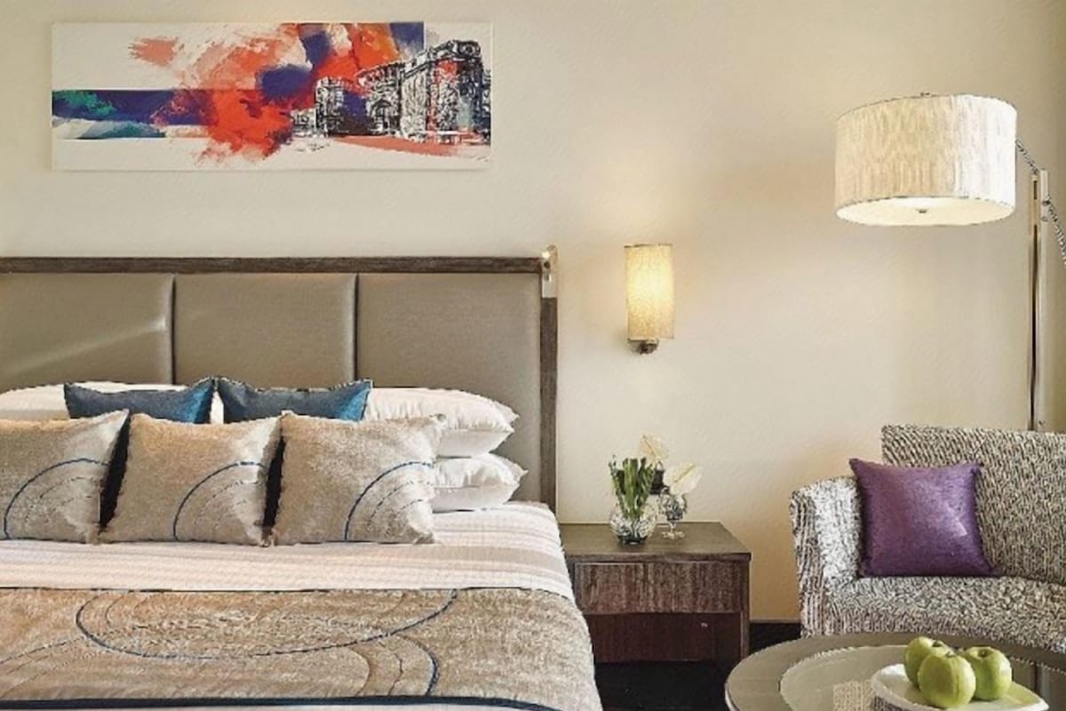 A hotel room with a bed, a nightstand, a lamp, a chair, and a painting on the wall.