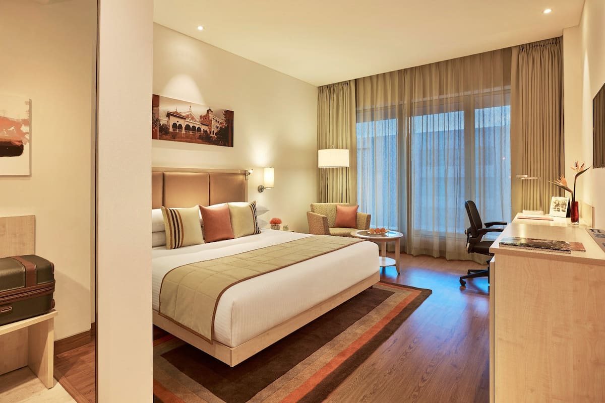 A hotel room with a large bed, a desk, a chair, a small table, and a window with curtains.