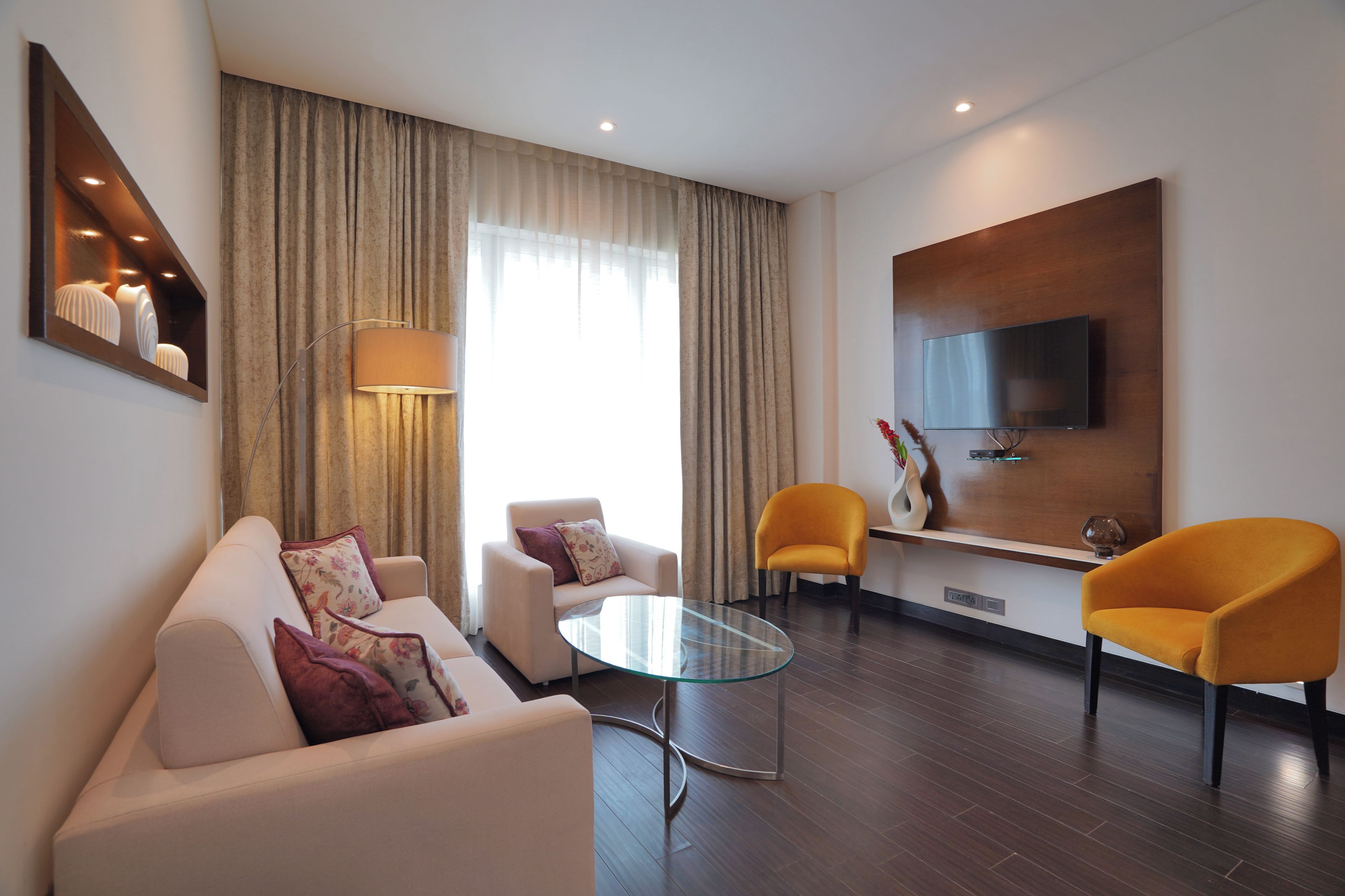 A modern living room with a sofa, armchairs, a glass coffee table, a flat-screen TV mounted on the wall, and a large window with curtains.