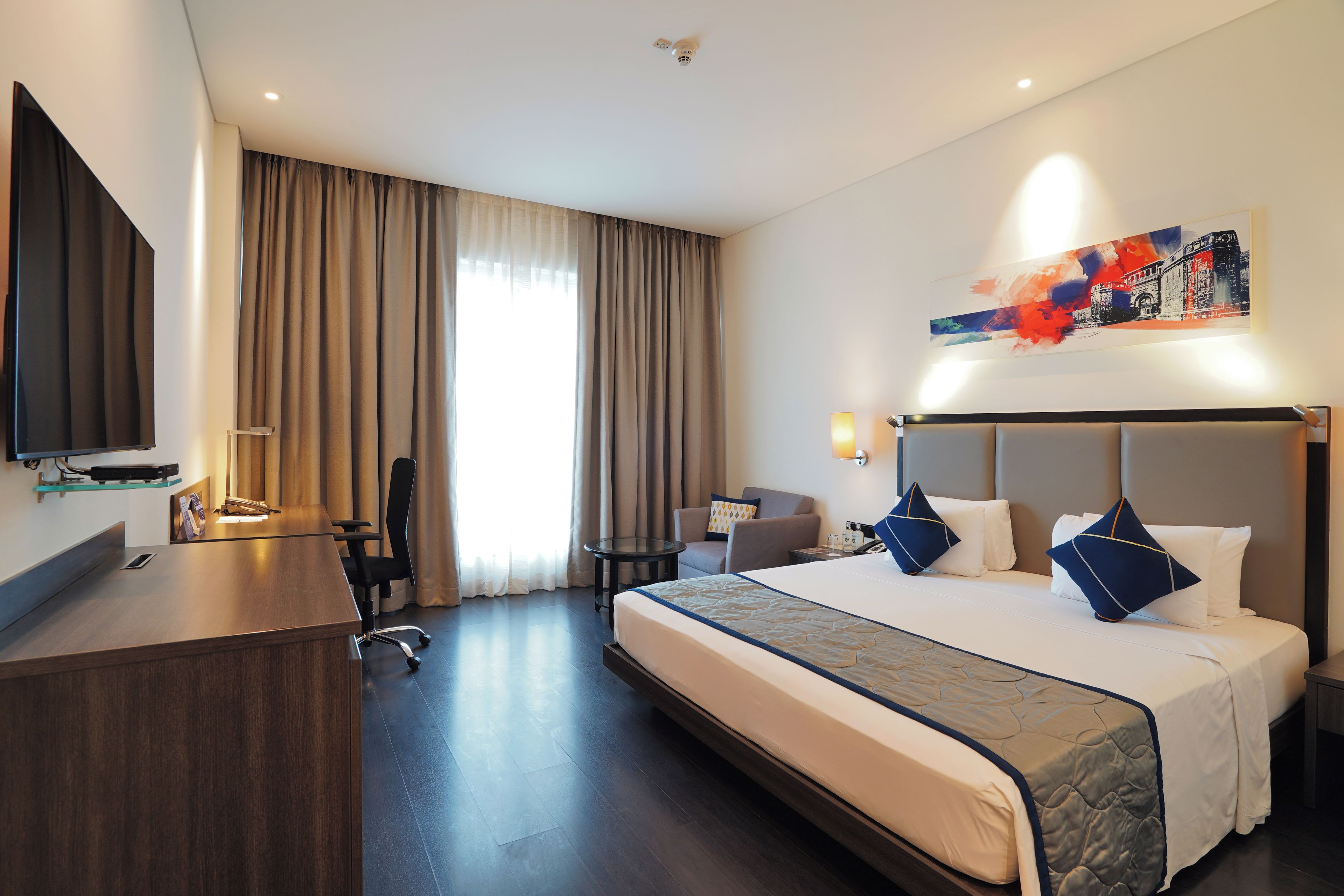 A hotel room with a large bed, a desk, a chair, a television, and a painting on the wall.