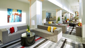 A spacious hotel lobby with a checkered floor, grey sofas with yellow and red cushions, a black coffee table, and a large abstract painting on the wall.