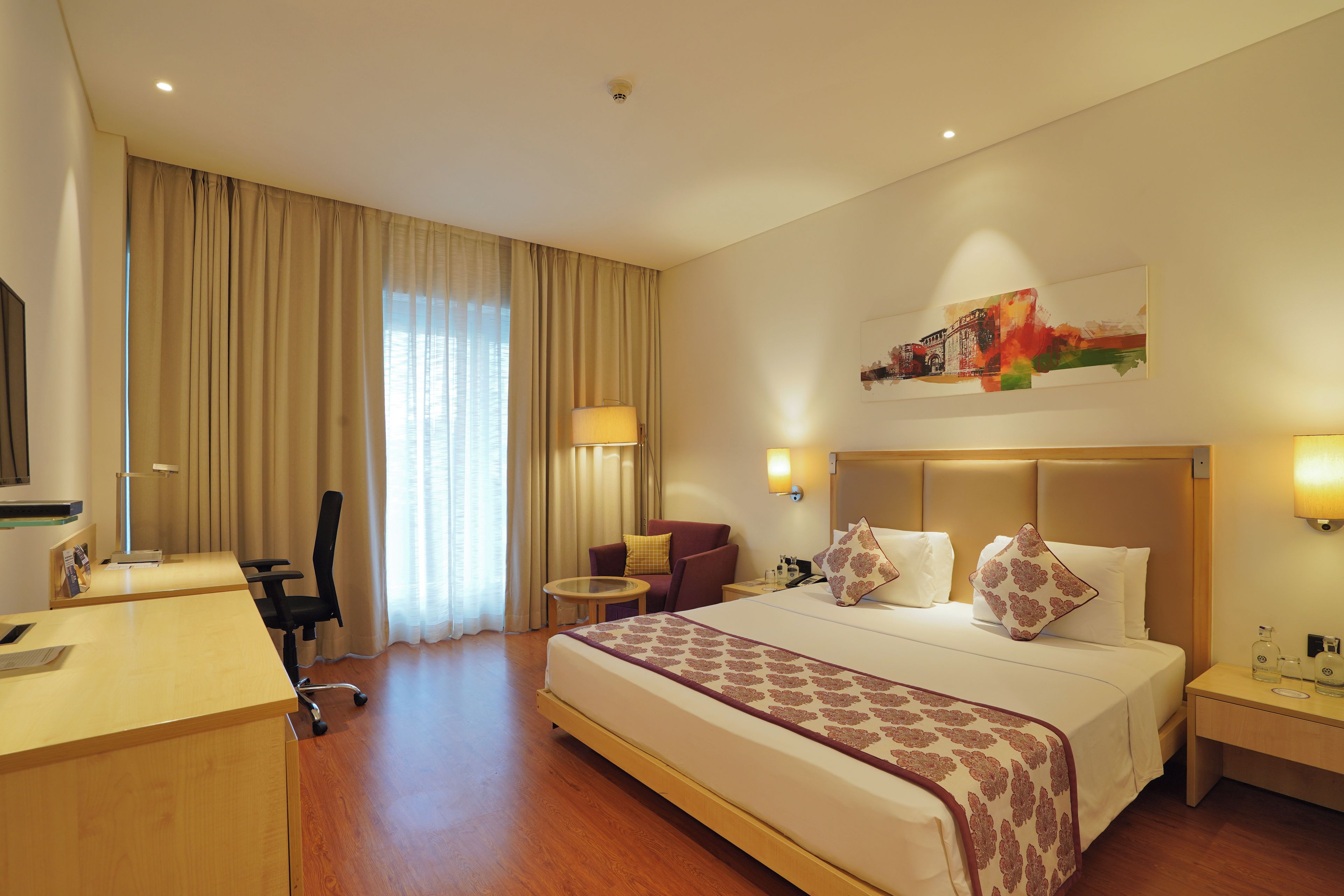 A hotel room with a bed, a desk, a chair, a small table, and a painting on the wall.