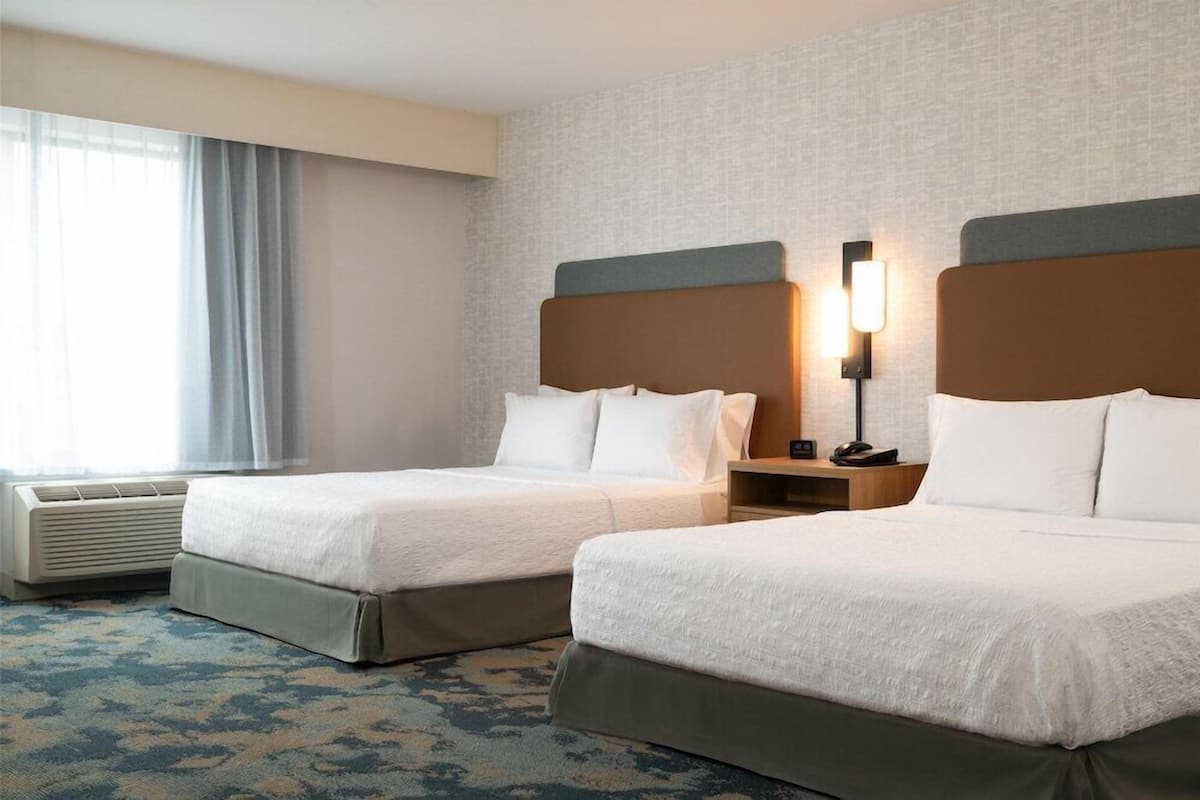 A hotel room with two beds, a bedside table with a lamp, a window with curtains, and a patterned carpet.