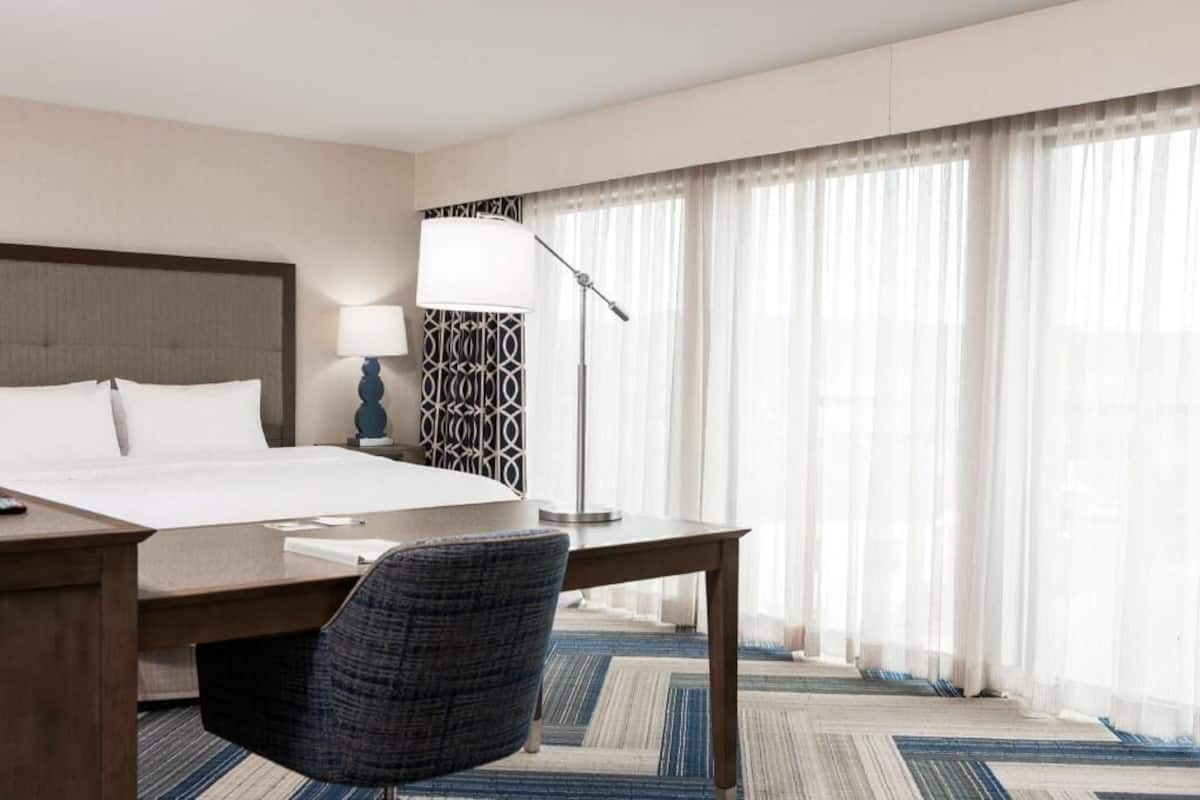 A hotel room with a bed, desk, chair, lamp, and large window with curtains.