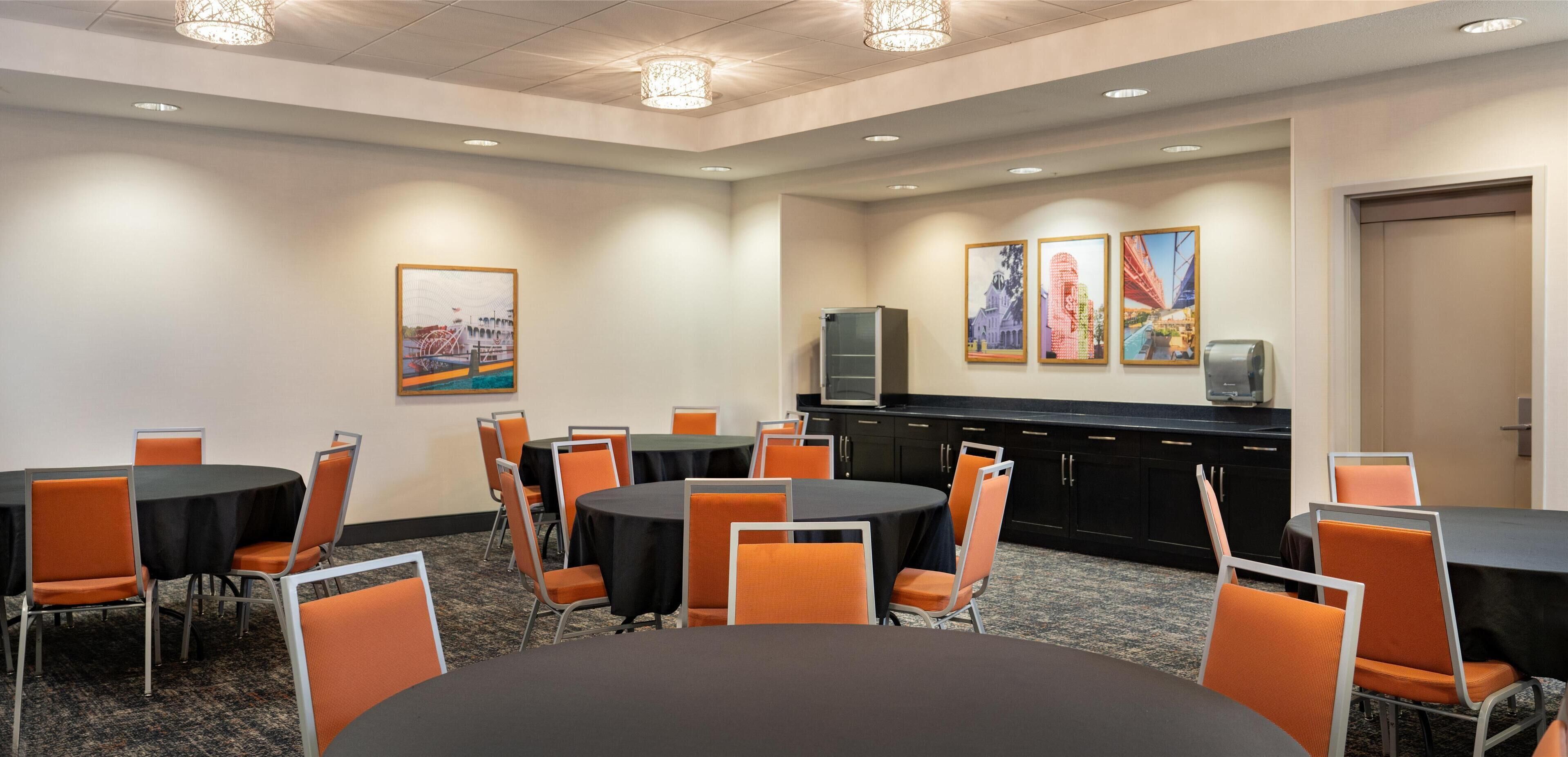 A conference room with round tables, orange chairs, a black cabinet, and framed pictures on the wall.