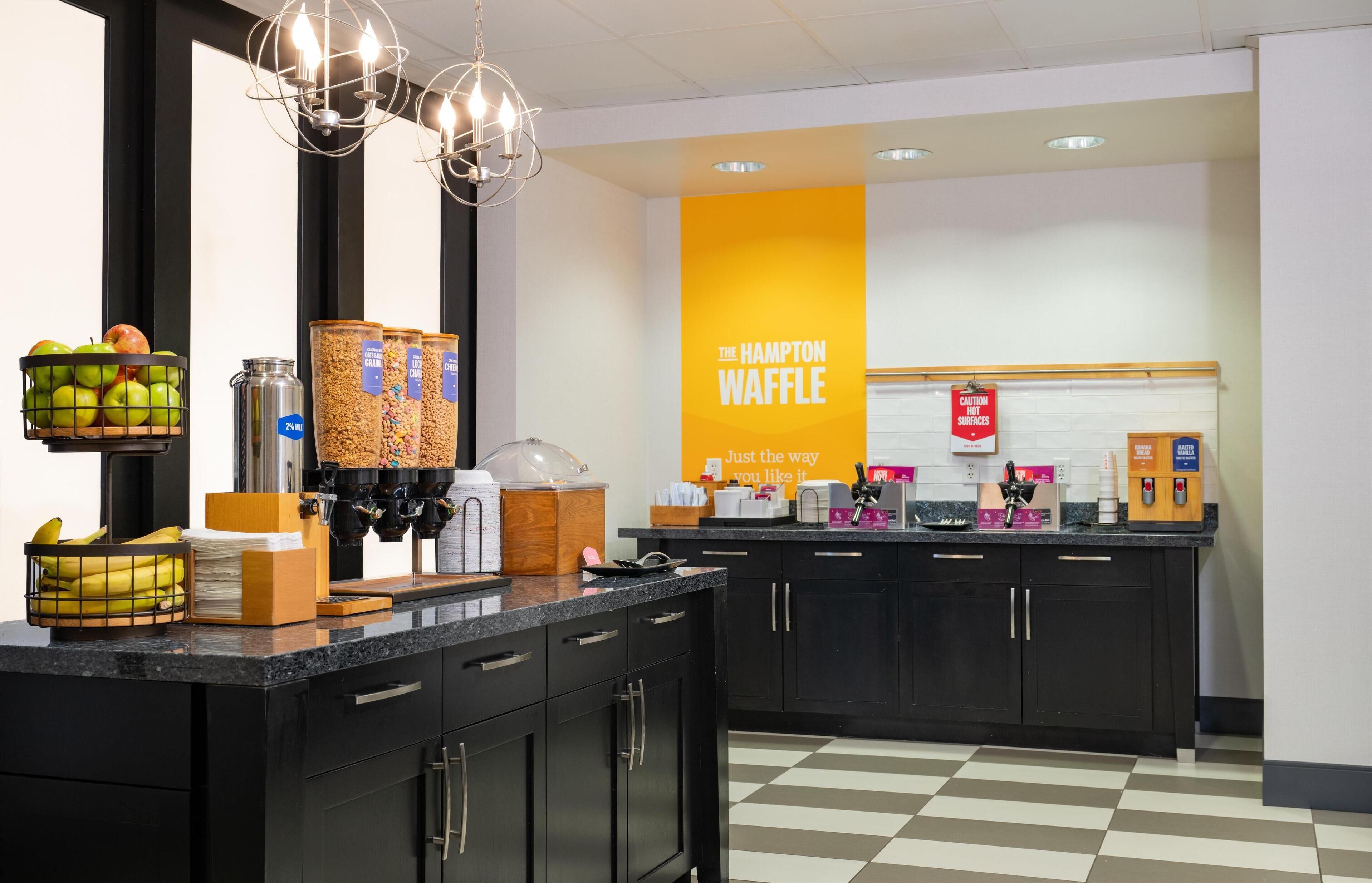 Uma área de cozinha com estação de waffles, cesta de frutas e máquina de café.