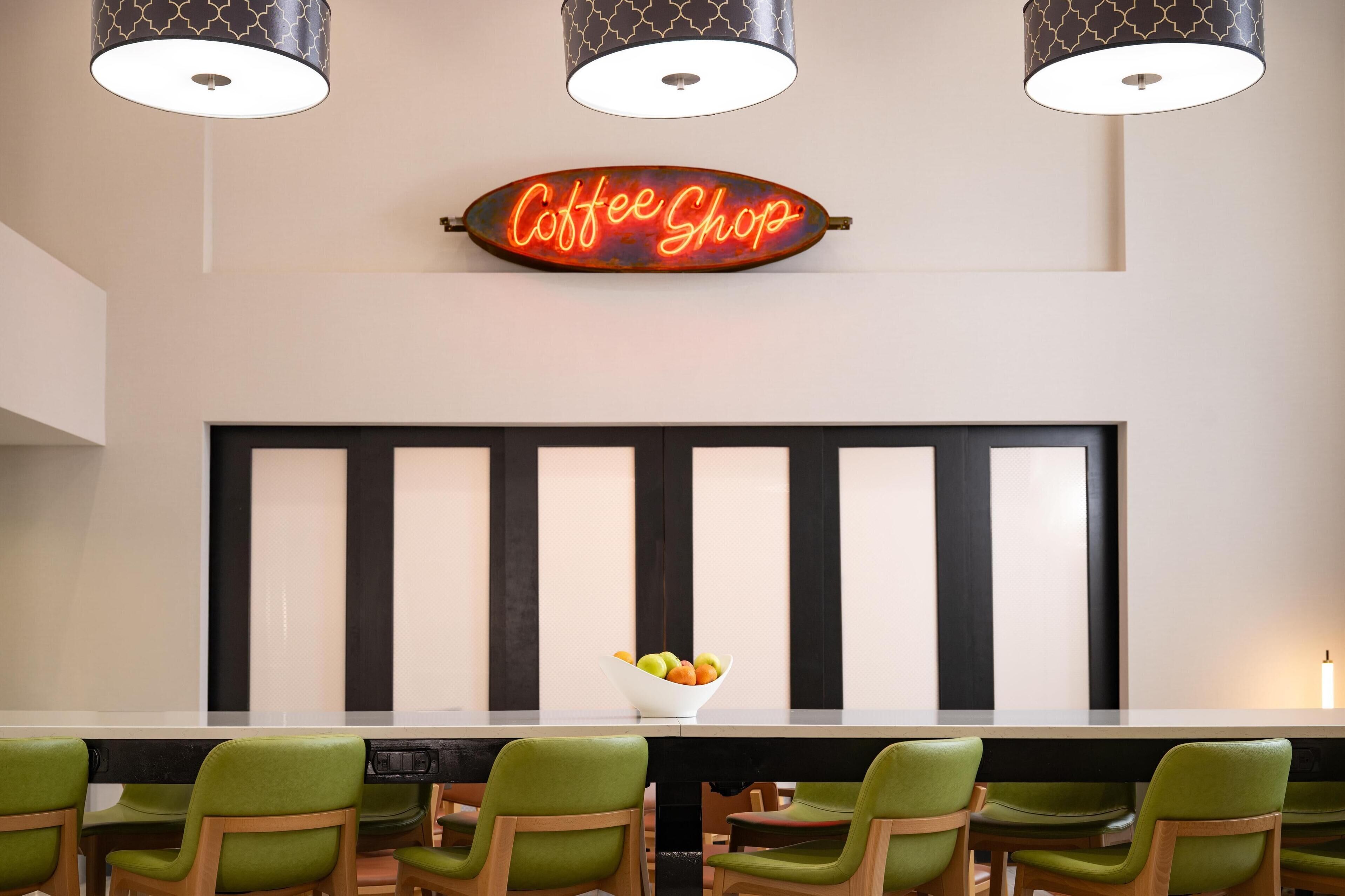 A modern coffee shop interior with a neon sign, a long table with chairs, and a fruit bowl.