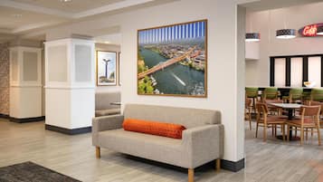 A modern hotel lobby with a large cityscape painting, a seating area with a sofa and chairs, and a dining area with tables and chairs.