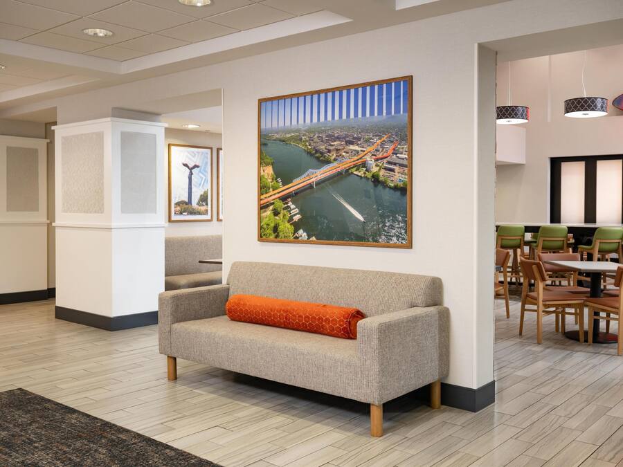 A modern hotel lobby with a large cityscape painting, a seating area with a sofa and chairs, and a dining area with tables and chairs.