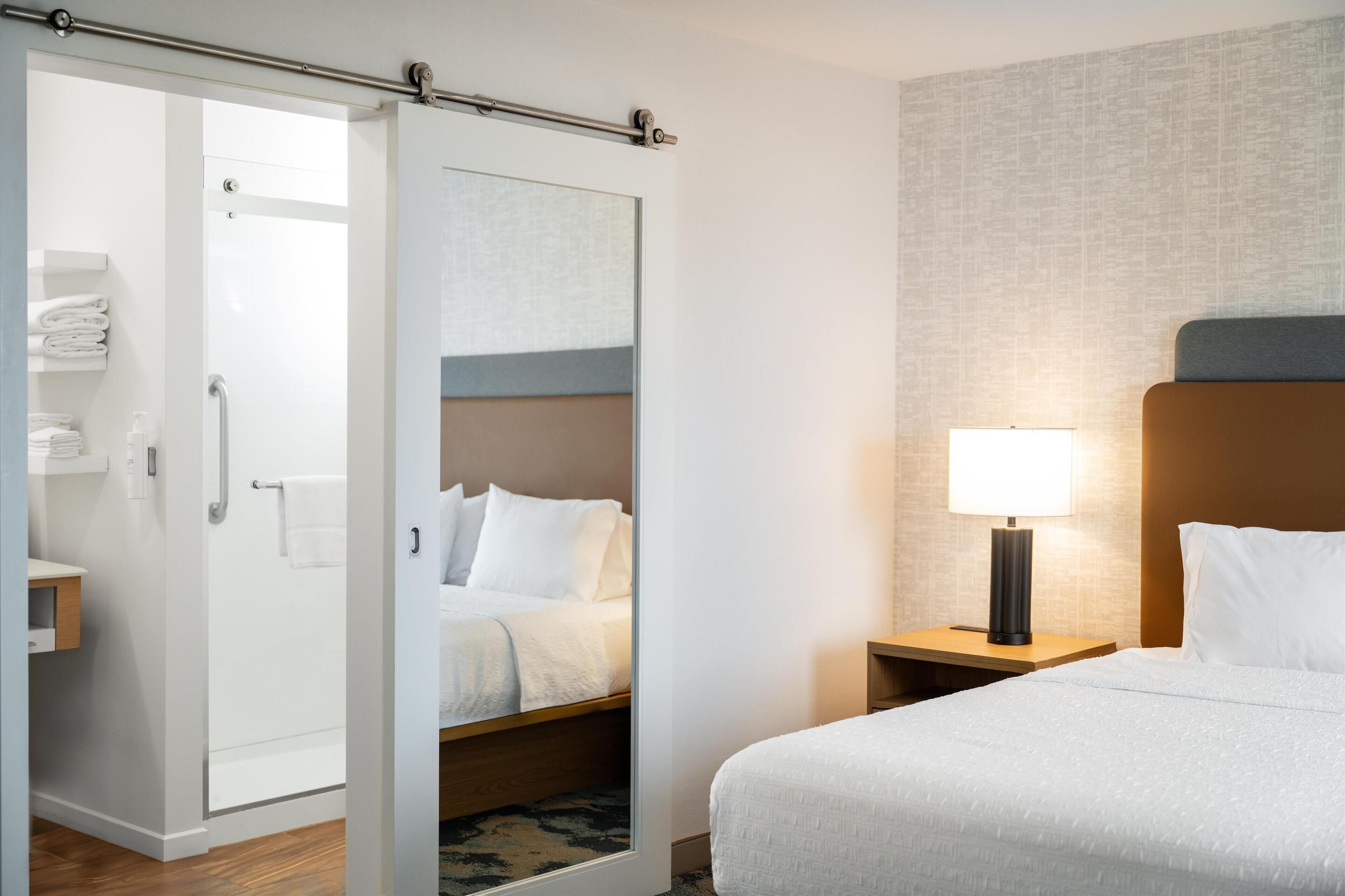 Une chambre d’hôtel comprenant un lit, une table de chevet avec une lampe, une salle de bain avec une douche et un miroir.