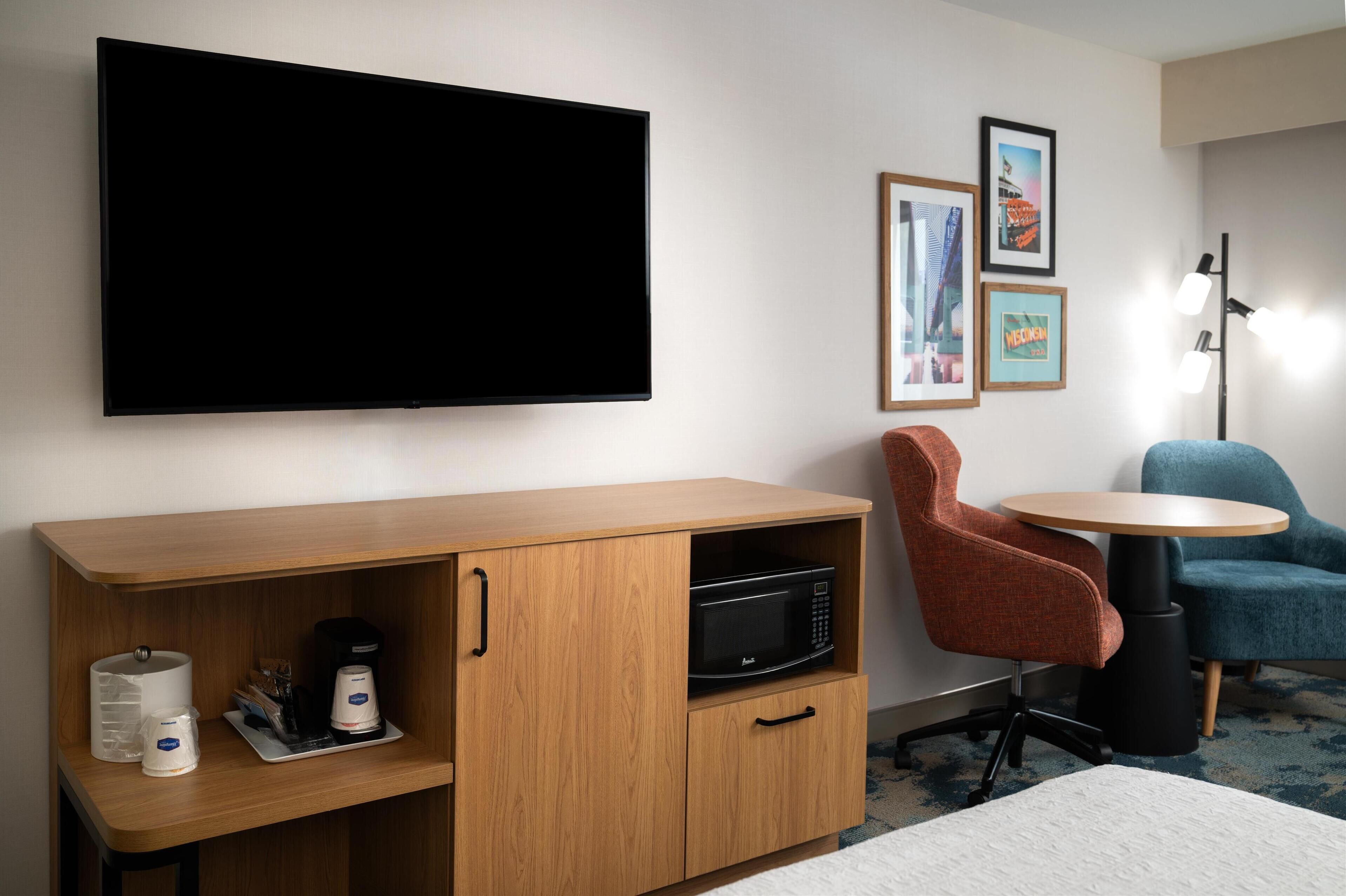 Ein Hotelzimmer mit Flachbildfernseher, einem Holfach mit Mikrowelle, einer Kaffeemaschine und einem kleinen Tisch mit zwei Stühlen.
