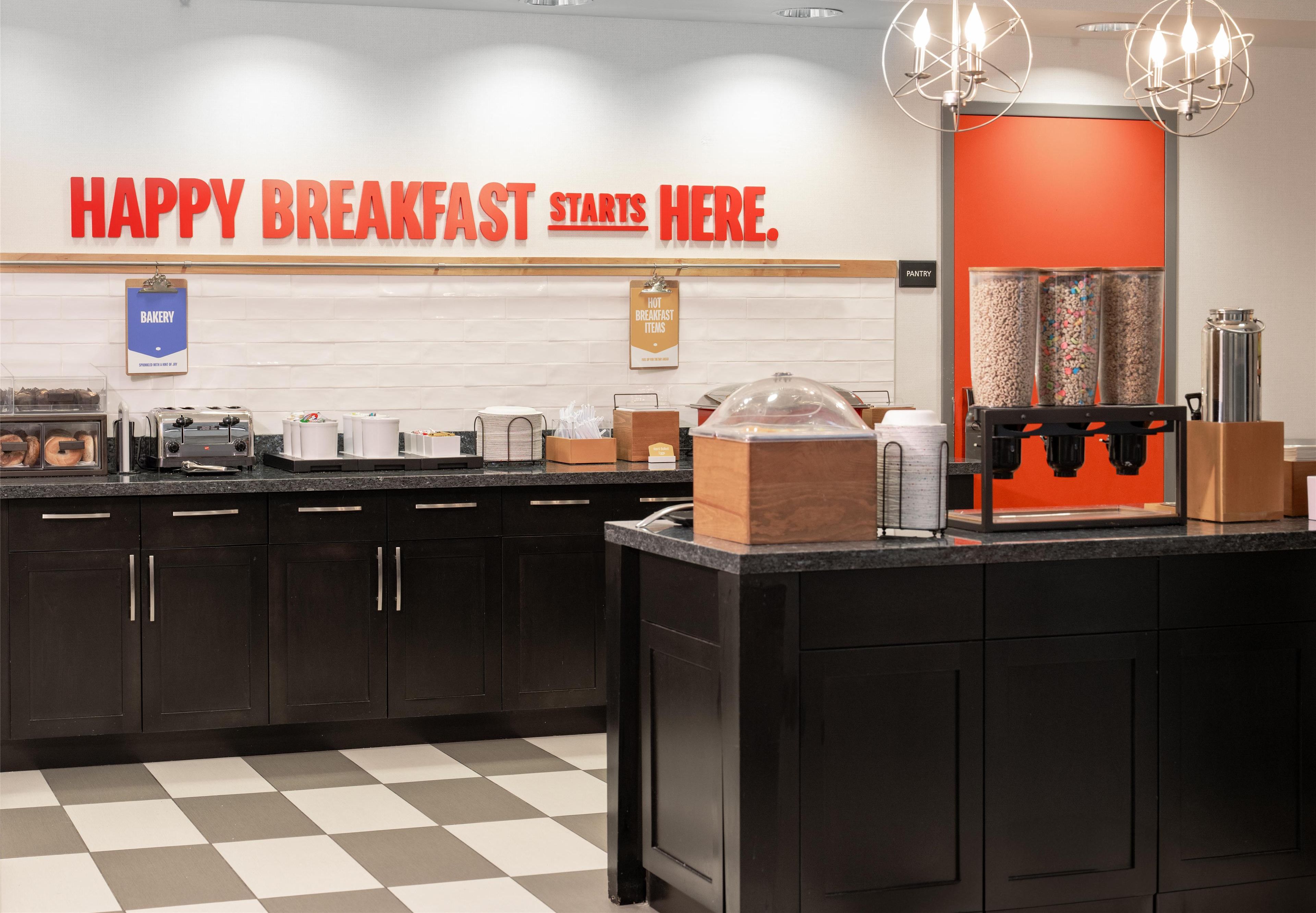 A breakfast area with a sign that reads "HAPPY BREAKFAST STARTS HERE." It includes a coffee machine, a food display, and a beverage dispenser.