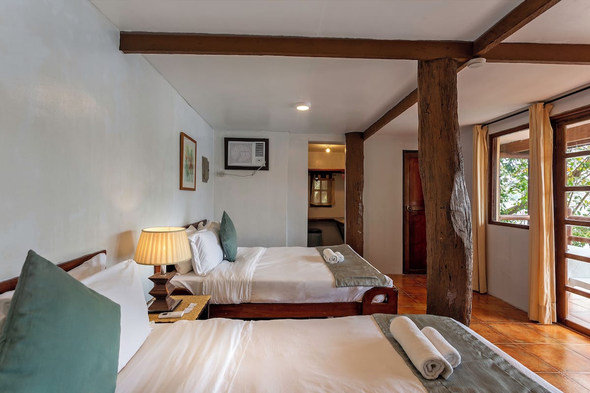 Een hotelkamer met twee bedden, houten balken, een plafondventilator en een raam met gordijnen.