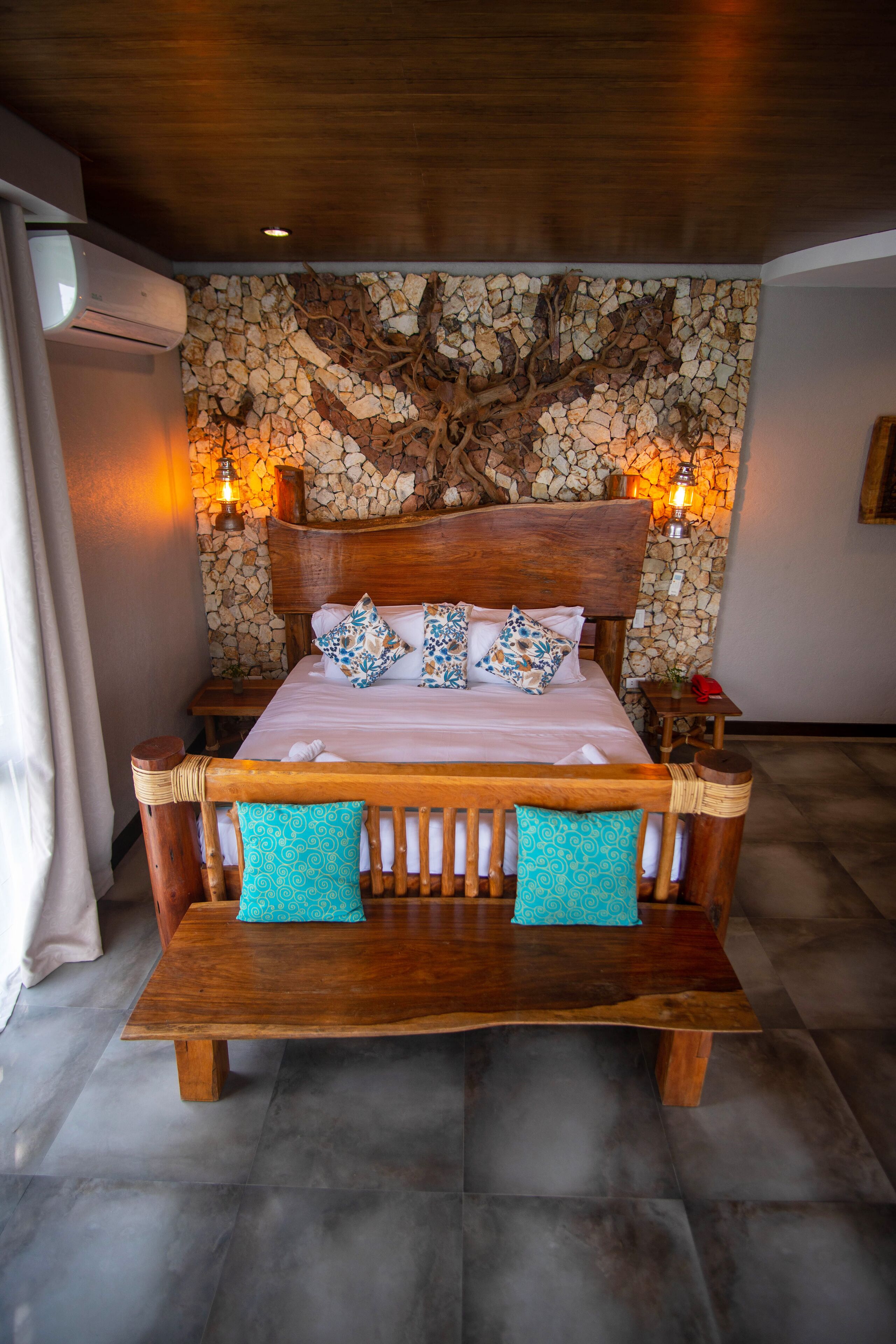 A wooden bed with a stone wall backdrop, two bedside lamps, and a wooden nightstand with a red object on it.