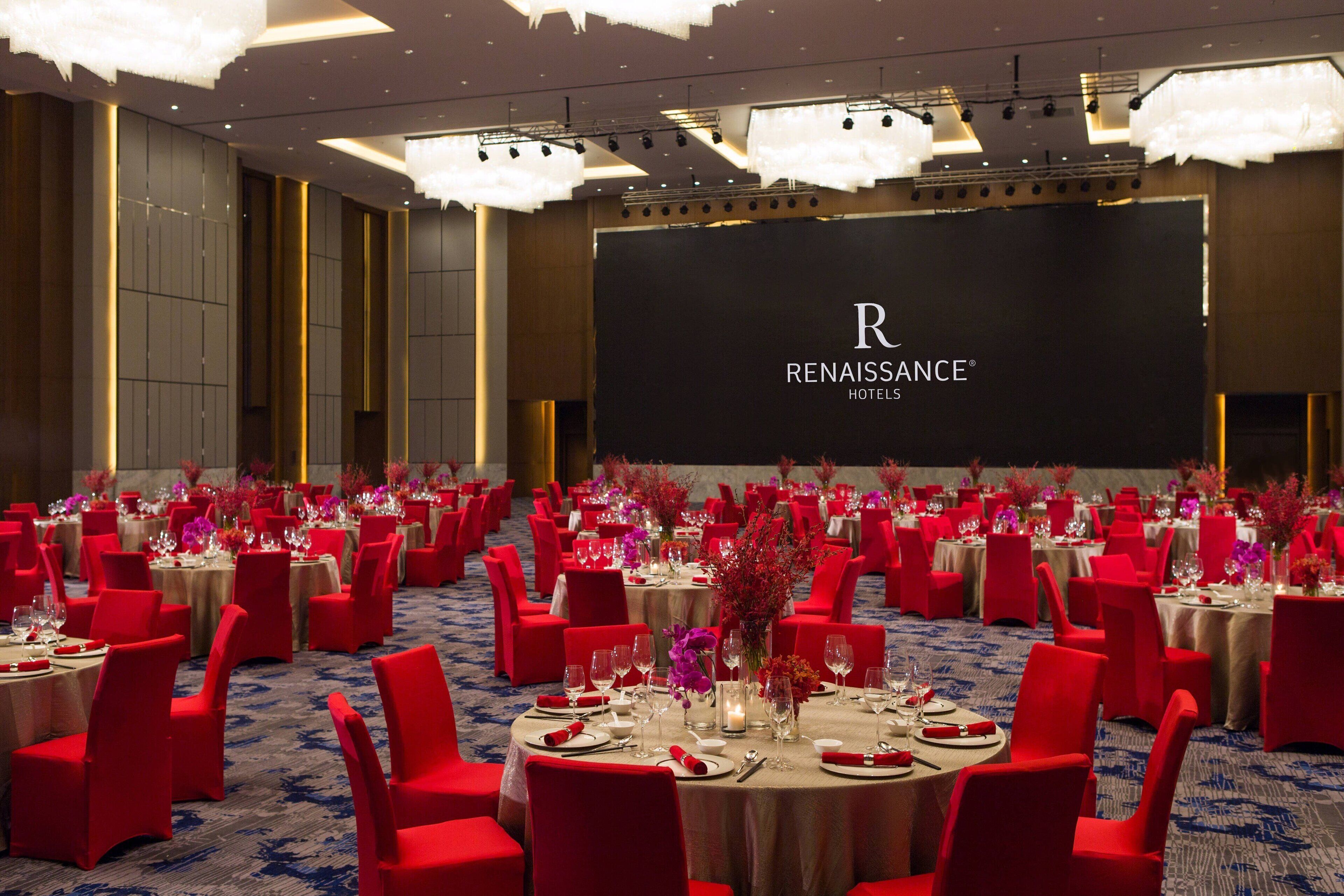 A banquet hall with round tables, red chairs, and a large screen displaying the RENAISSANCE HOTELS logo.