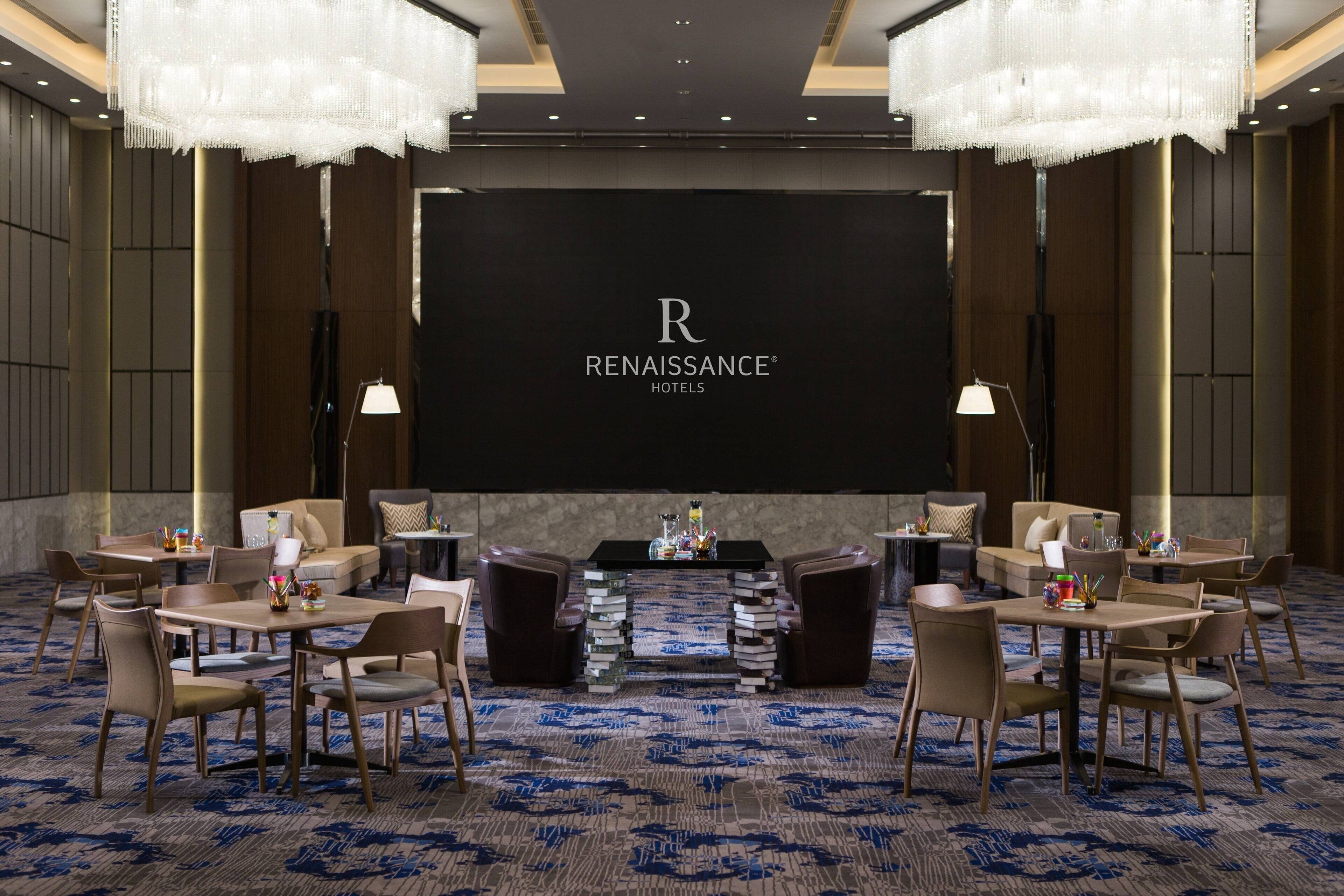 Un salón de conferencias con una pantalla grande mostrando el logotipo de Renaissance Hotels. La sala tiene un diseño moderno con una alfombra estampada, varias mesas con sillas y lámparas decorativas.