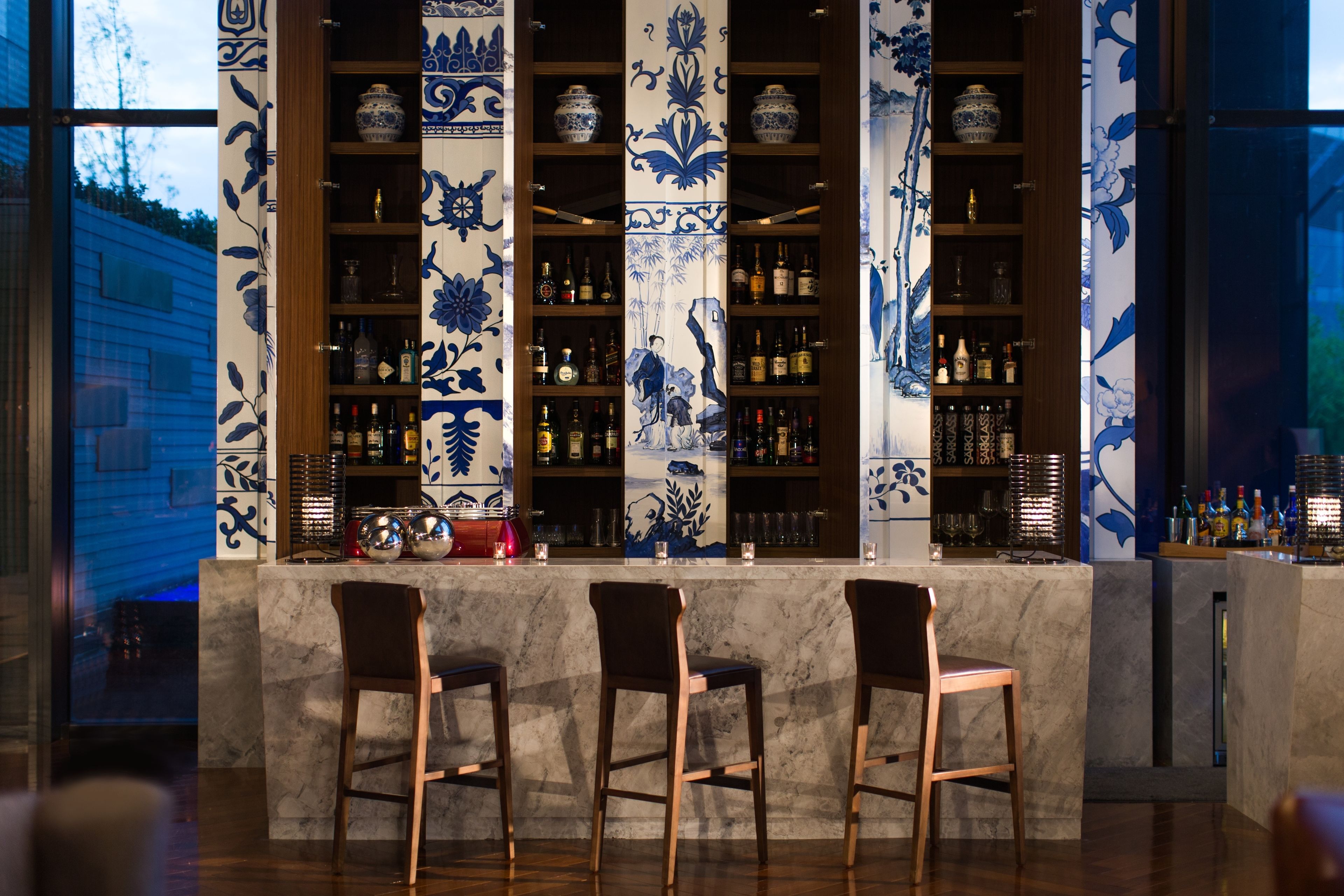 Un área de bar con tres taburetes de madera, estantes con botellas y azulejos decorativos azules y blancos.