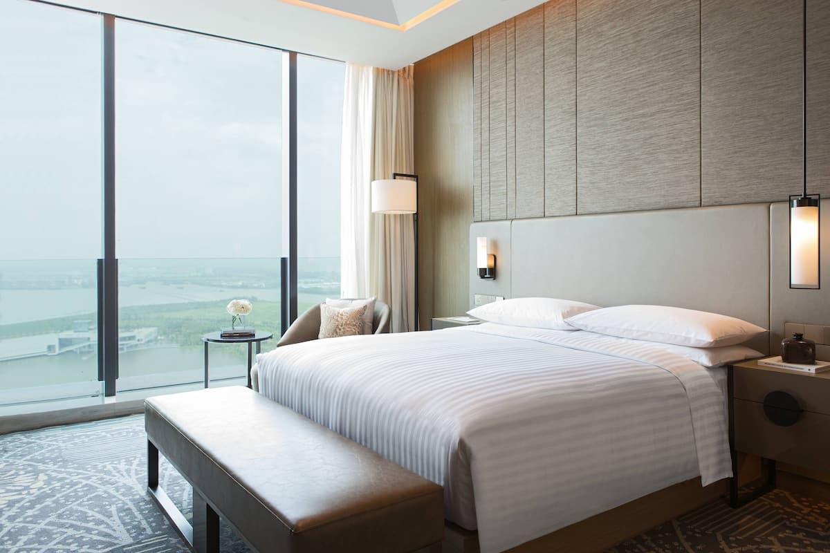 A modern hotel room with a large bed, a bench, a nightstand, and a view of the city.