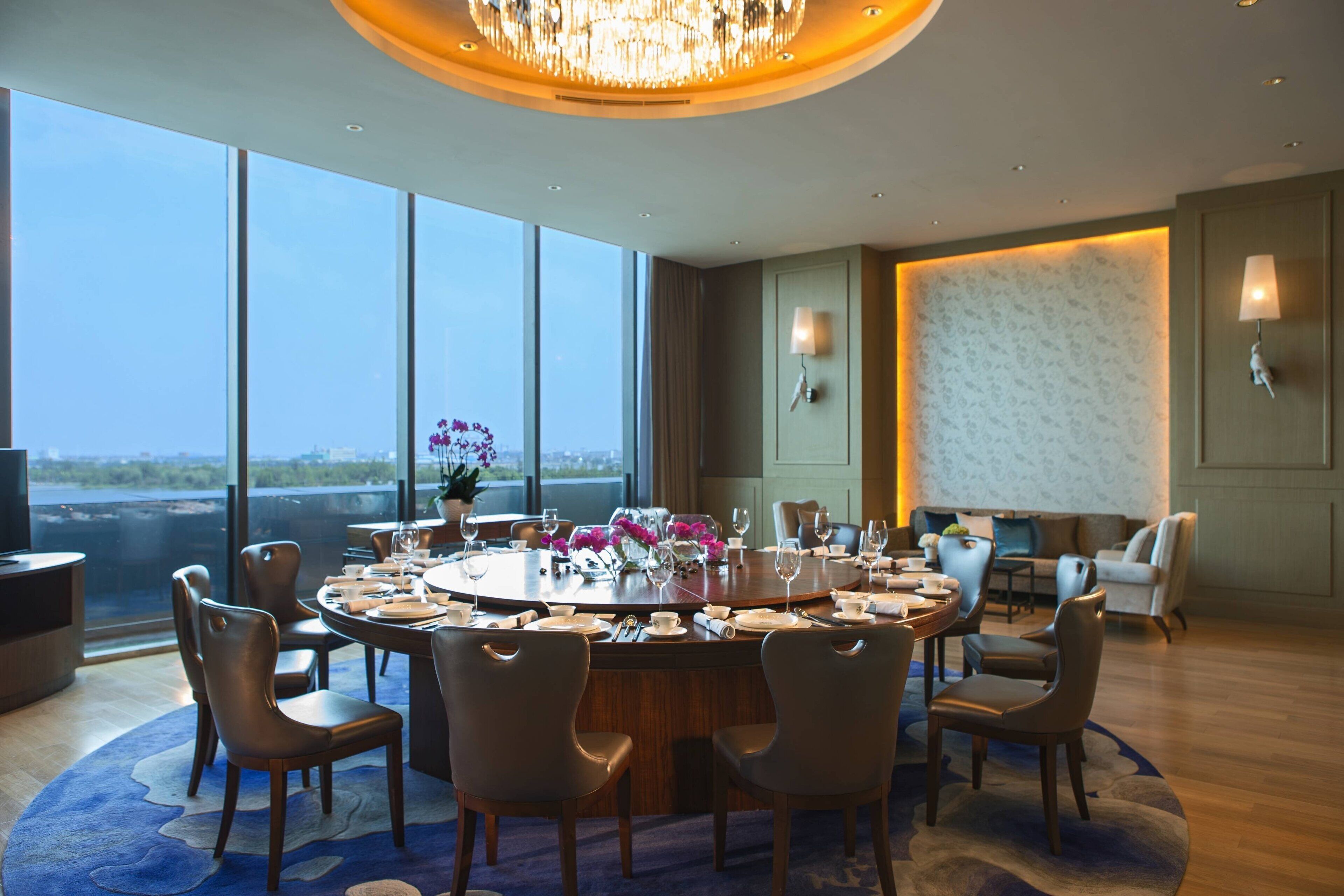 A spacious dining area with a round table set for six, surrounded by chairs, large windows offering a city view, and a chandelier above.