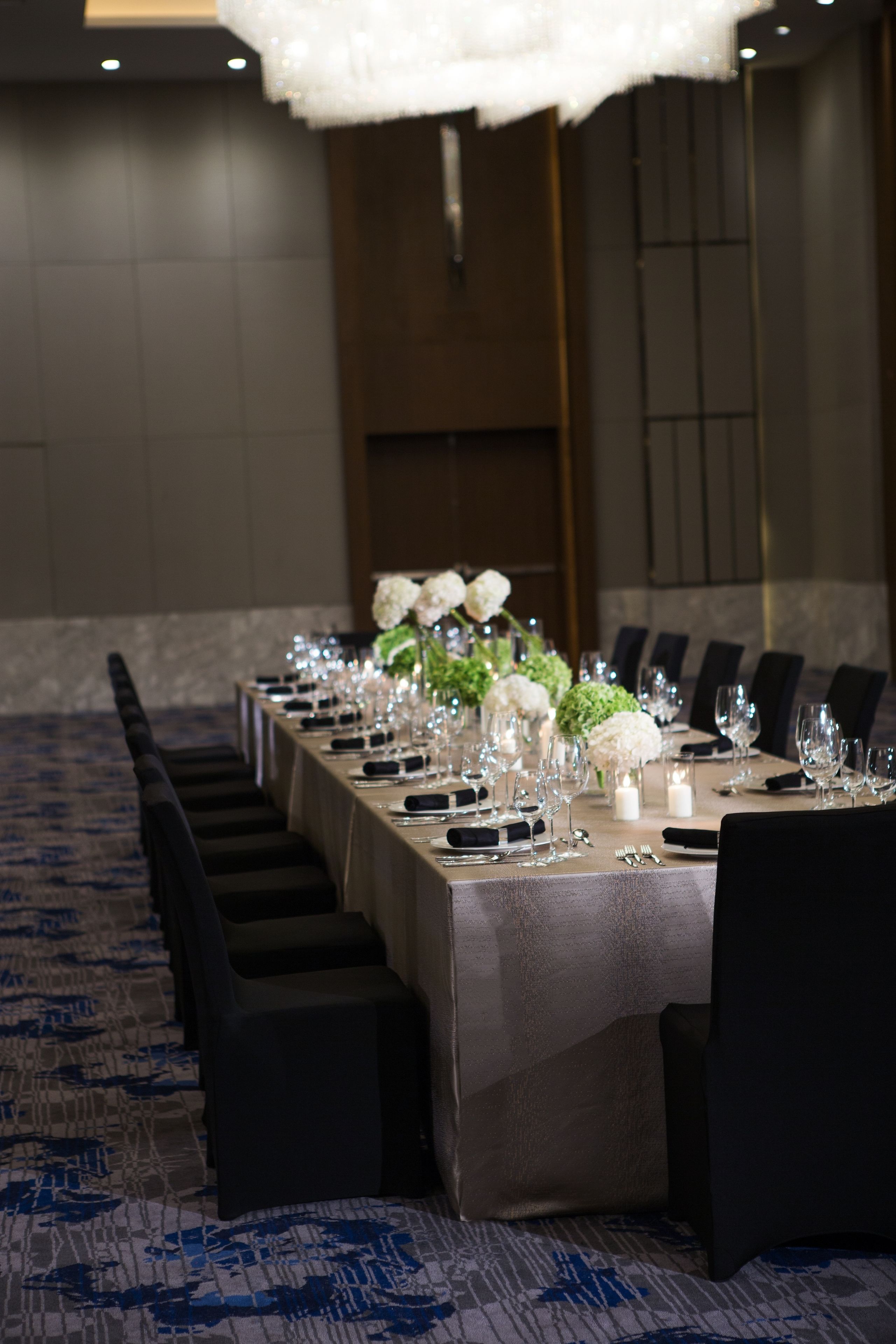 A long banquet table set with chairs, white flowers, and glassware in a room with a patterned carpet and a chandelier.