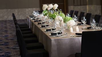 Una larga mesa banquete dispuesta con sillas, flores blancas y cristalería en una habitación con alfombra estampada y un candelabro.