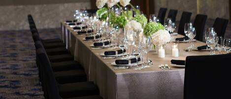 A long banquet table set with chairs, white flowers, and glassware in a room with a patterned carpet and a chandelier.