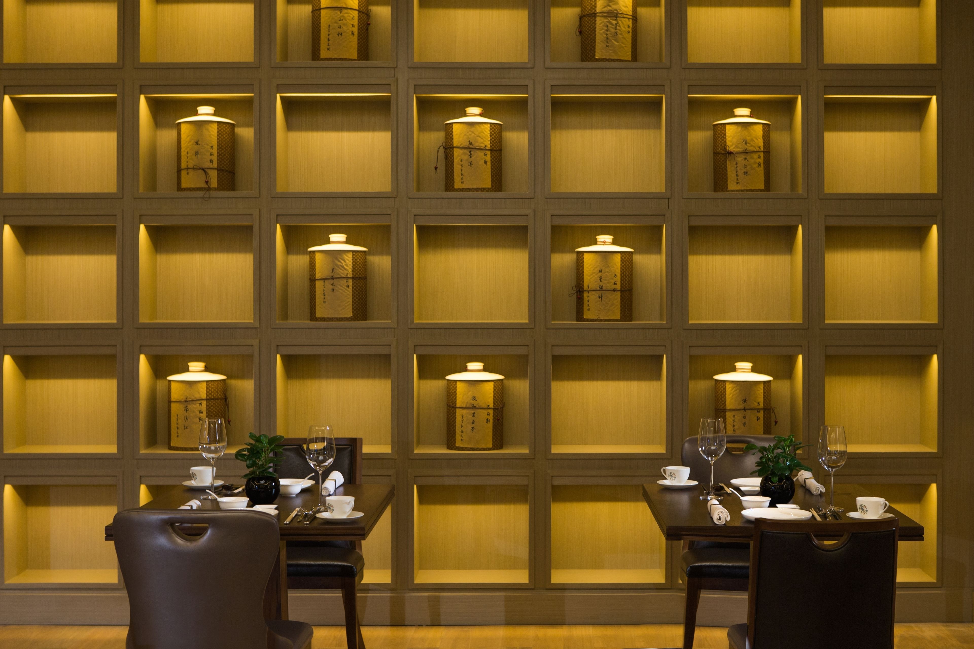 A dining area with a table set for two, surrounded by chairs, with a wall of illuminated shelves behind.