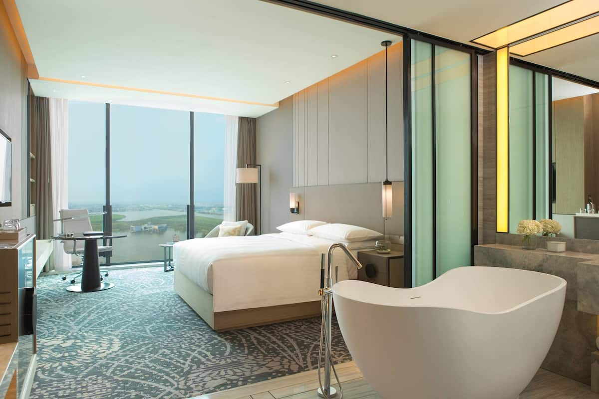 A modern hotel room with a large bed, a sitting area with a desk, a bathtub, and a view of the city.