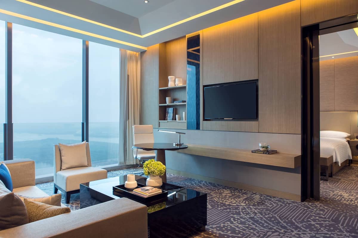 A modern hotel room with a large window, a flat-screen TV, a sofa, a coffee table, and a bed.